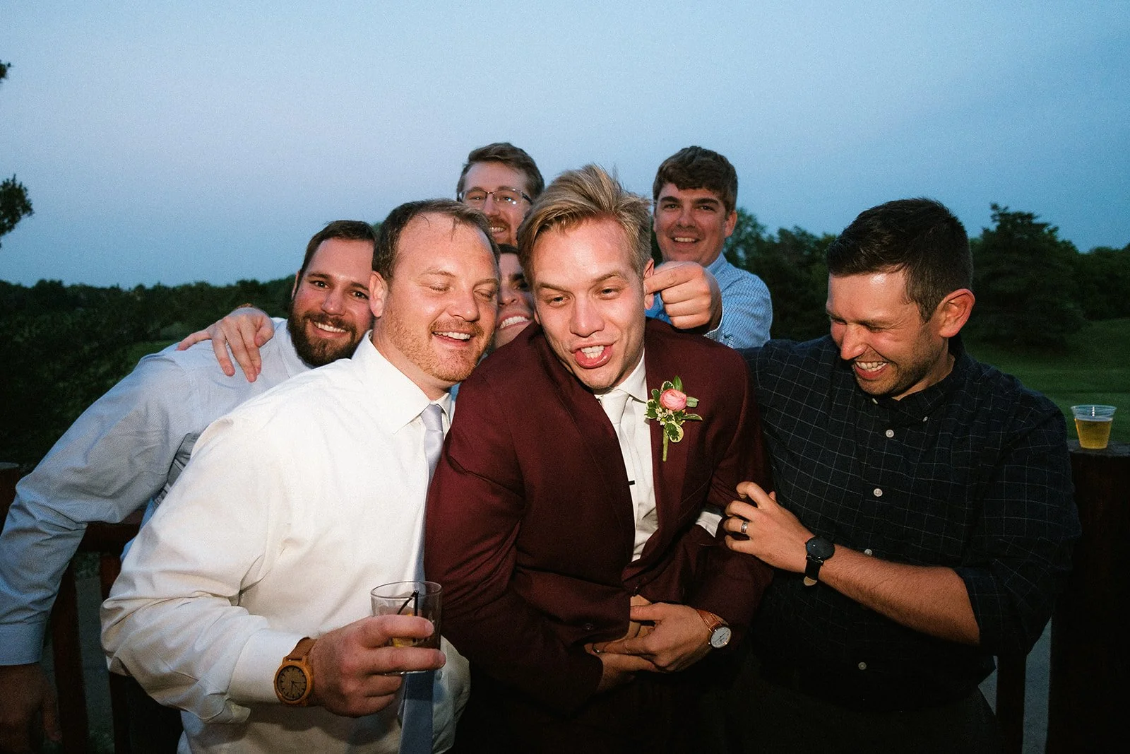 Group of seven men at an outdoor gathering, one of whom is wearing a suit with a boutonniere, smiling and joking with each other during evening.