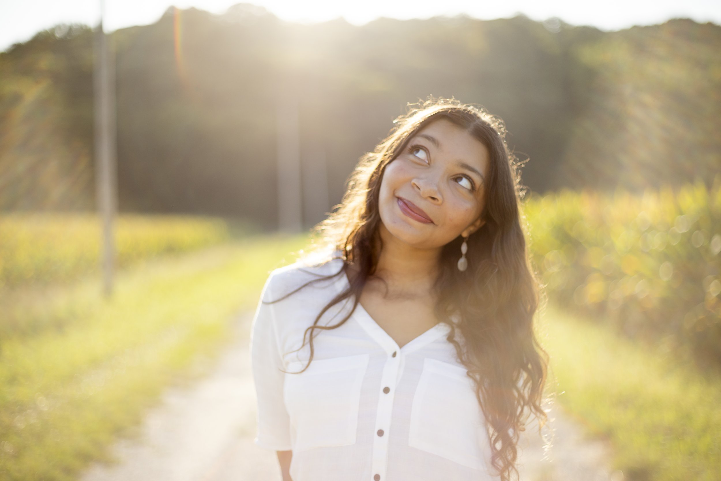 A young woman with long brown hair, wearing a white button-up shirt, standing outdoors in sunlight with a blurred natural background.