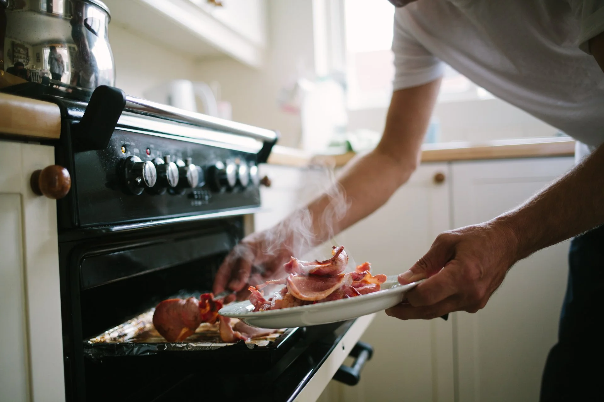 Person placing raw bacon and vegetables on a baking sheet in front of an oven in a kitchen.