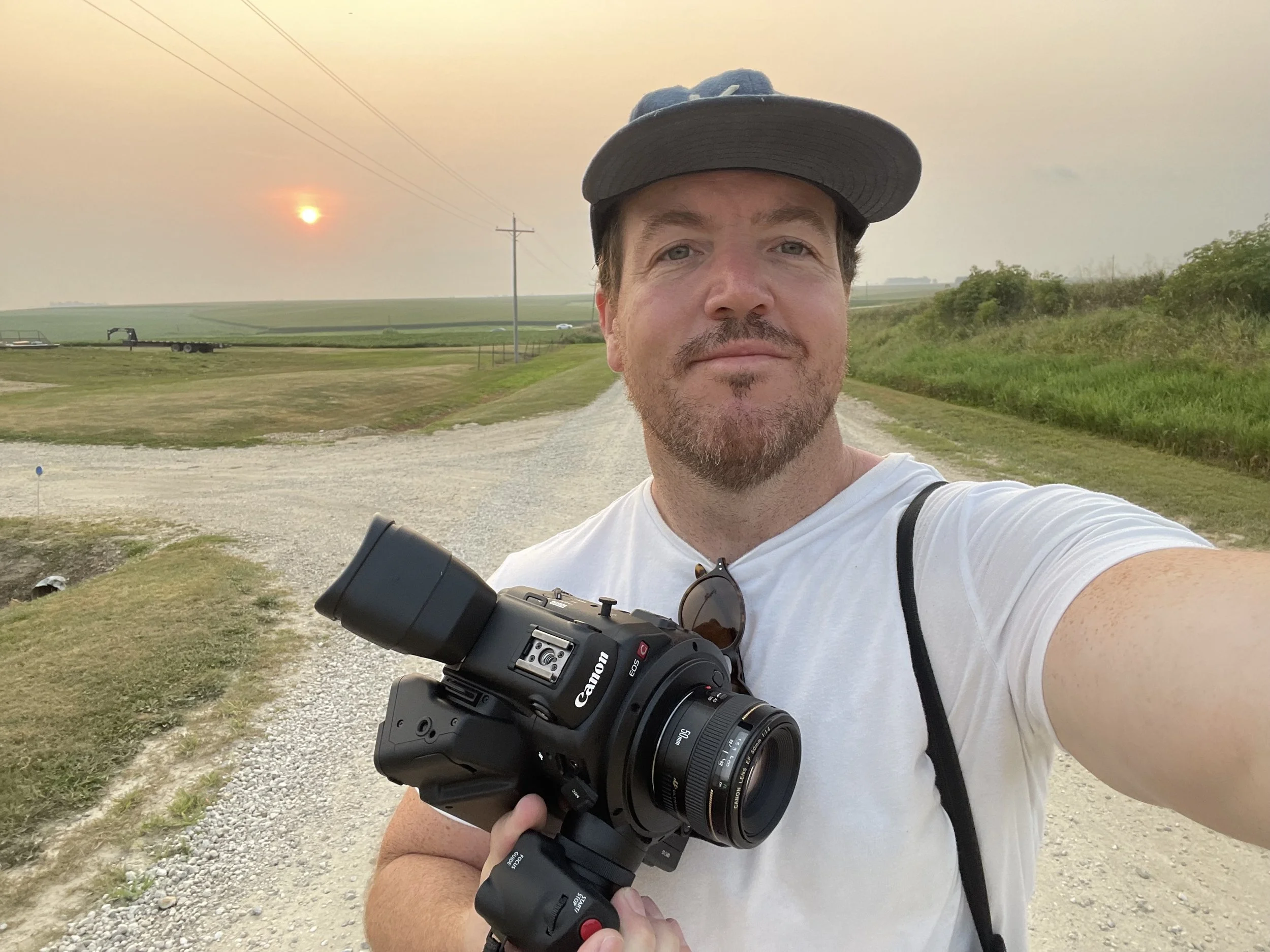 A man taking a selfie outdoors during sunset, holding a camera in his right hand, wearing a white shirt, gray cap, and sunglasses hanging on his shirt. A grassy path, green fields, and power lines are visible in the background.
