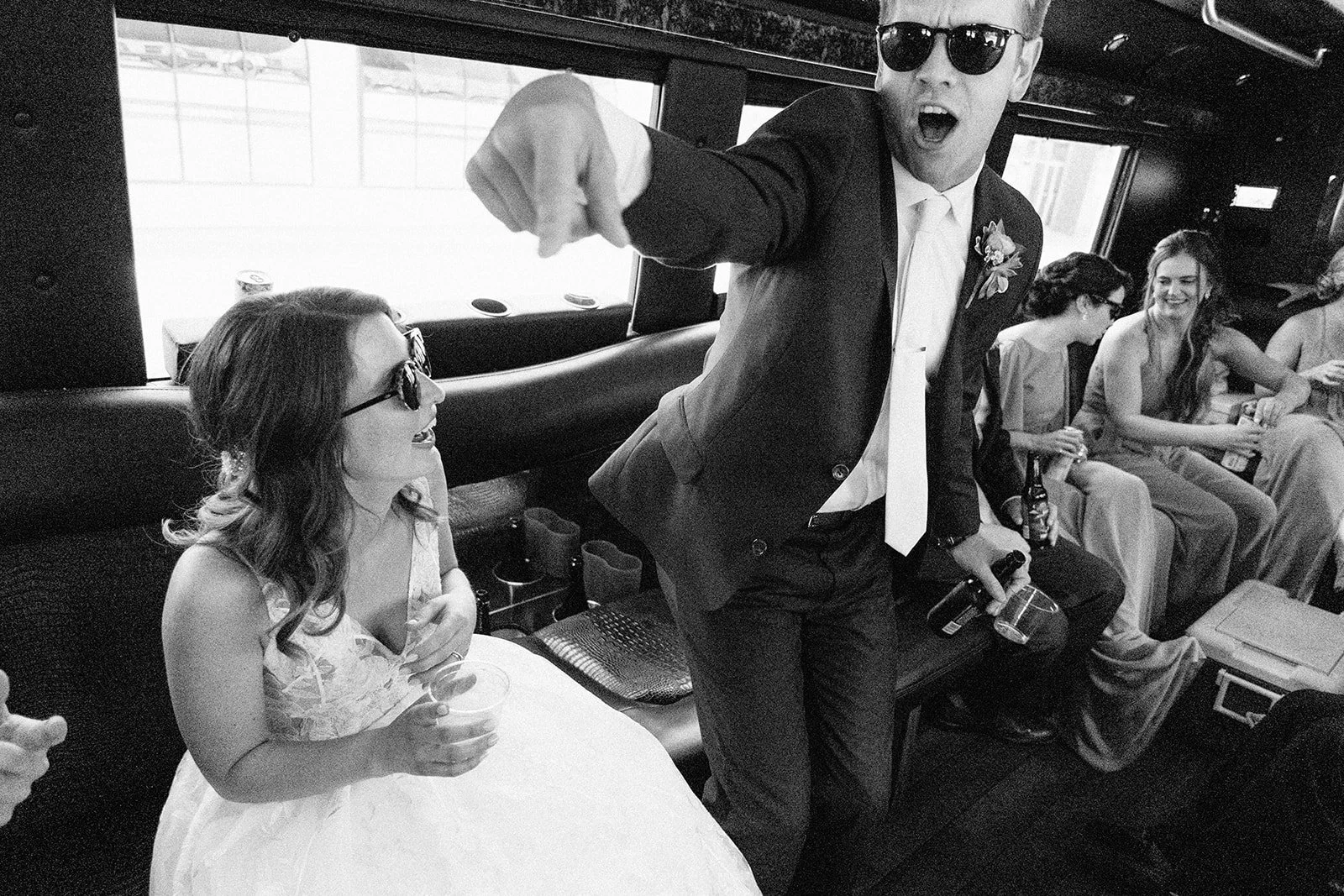 A man in a suit and sunglasses is pointing and shouting at a woman in a wedding dress, sitting and smiling, on a bus with other women sitting nearby, holding drinks.