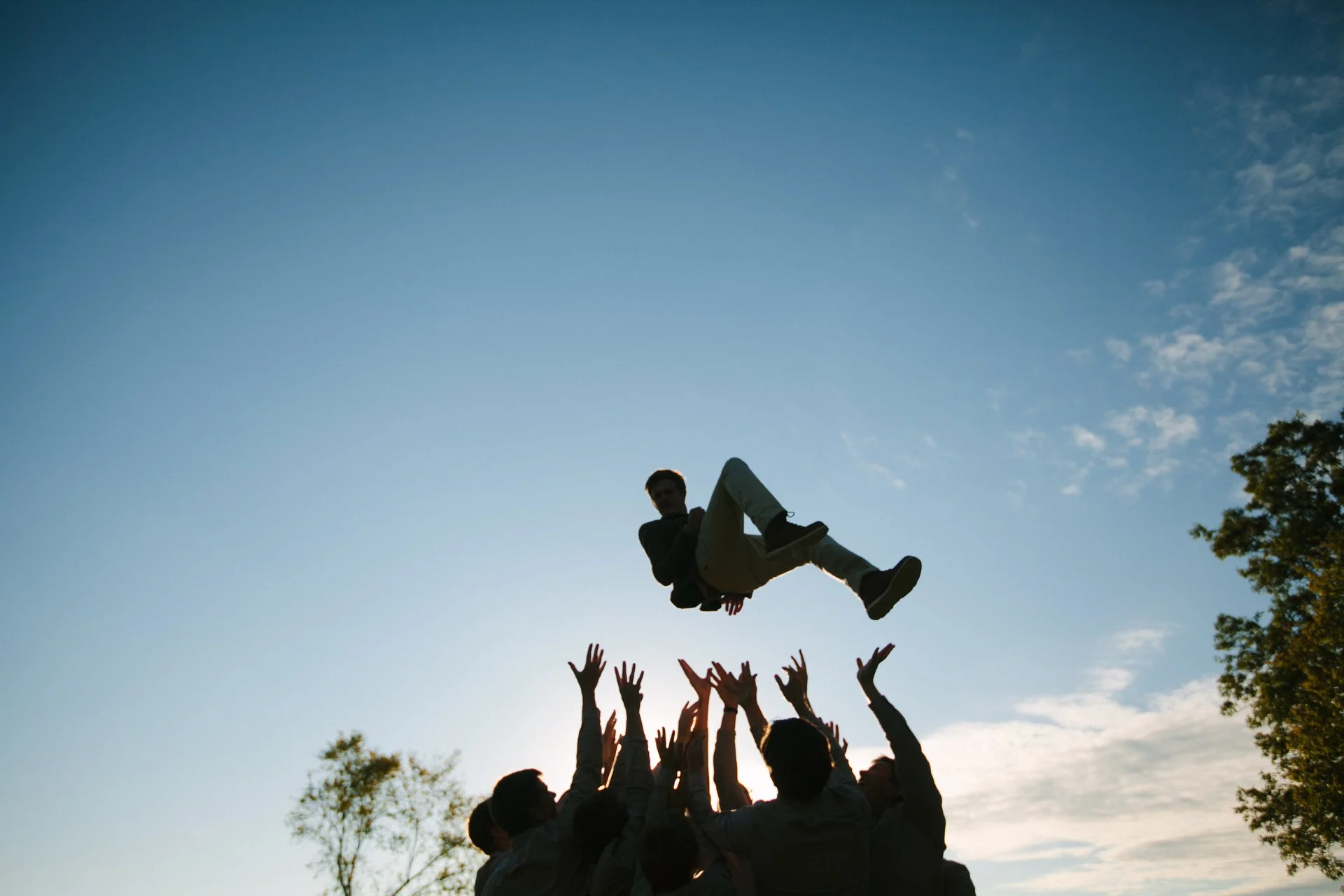 People forming a circle lifting a person into the air outdoors during sunset, with a clear sky and trees visible.
