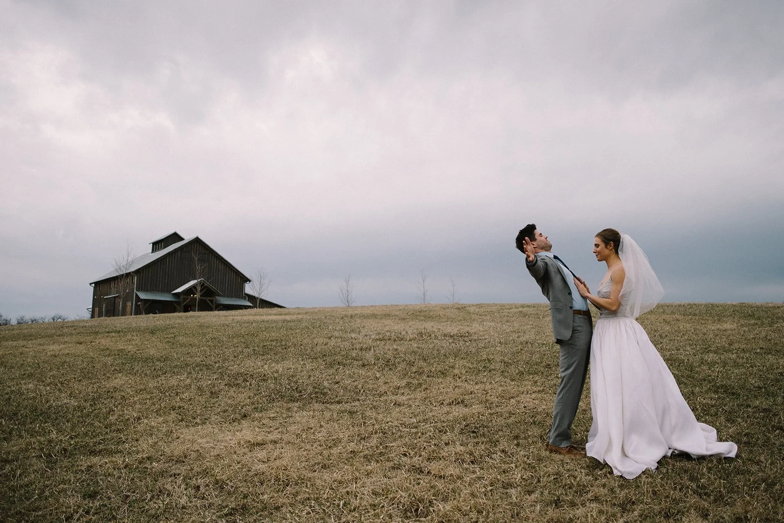 A bride and groom standing on a grassy hill outside under cloudy sky, with a rustic barn in the background, during their wedding photoshoot.
