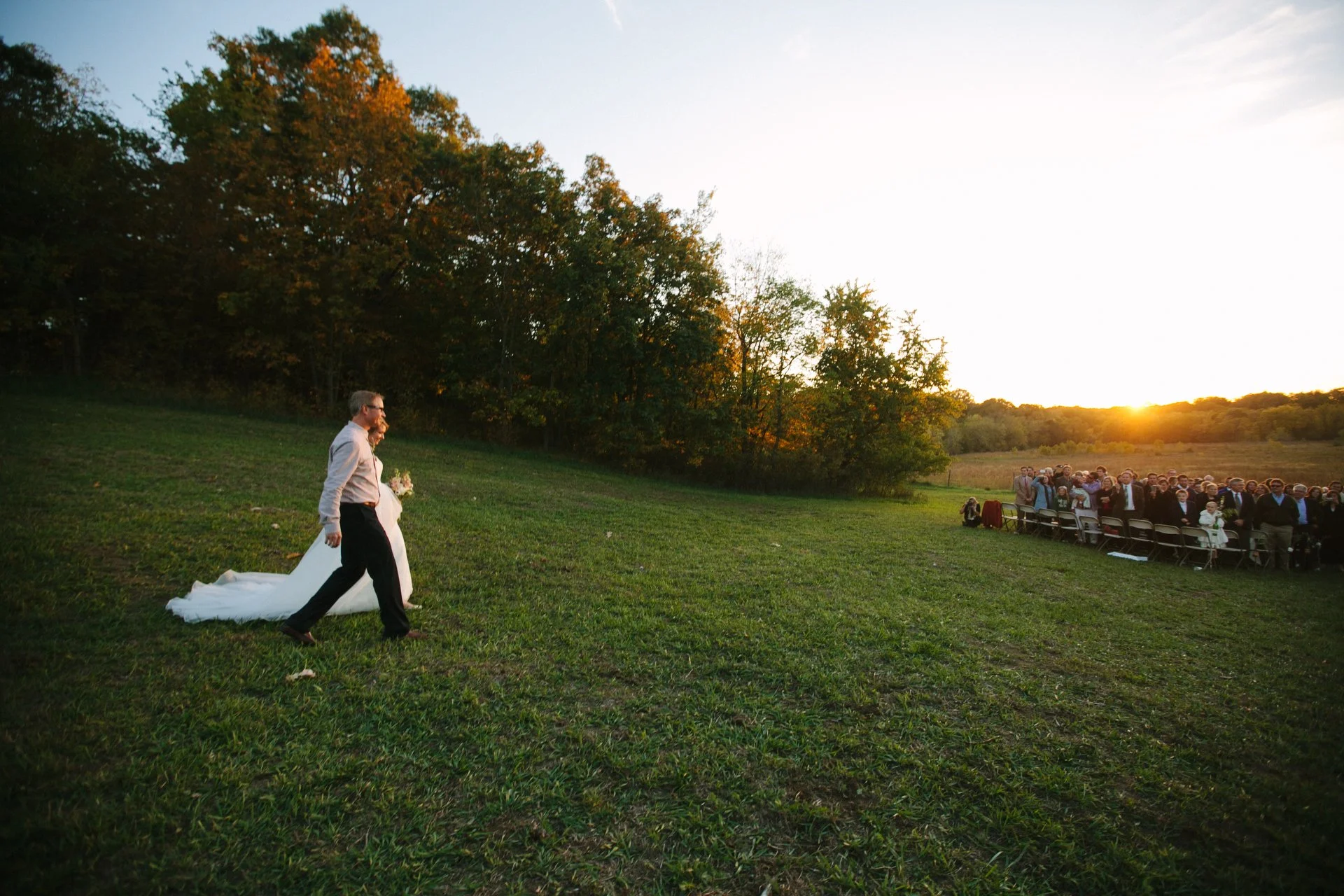 A wedding ceremony outdoors during sunset, with a bride and groom walking across a grassy field and guests seated in chairs watching, surrounded by trees with autumn foliage.