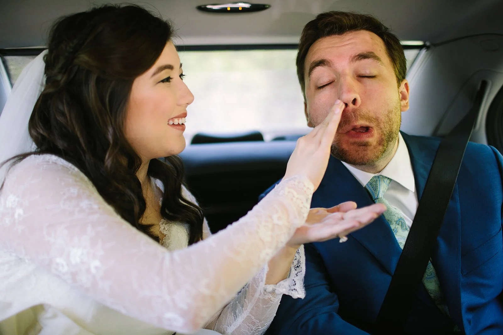 A woman in a wedding dress touches a man's nose while sitting in the back of a car. The man, dressed in a blue suit, appears sleepy or unwell.