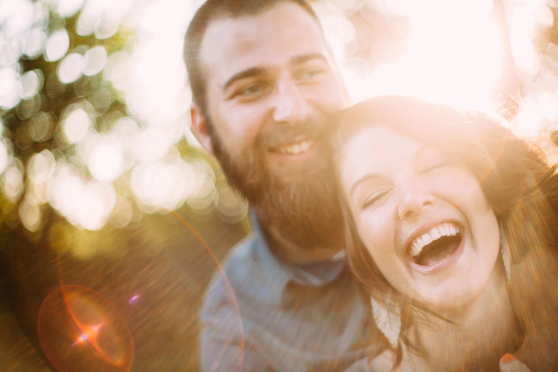 A man and woman smiling and laughing outdoors in sunlight with bokeh background.