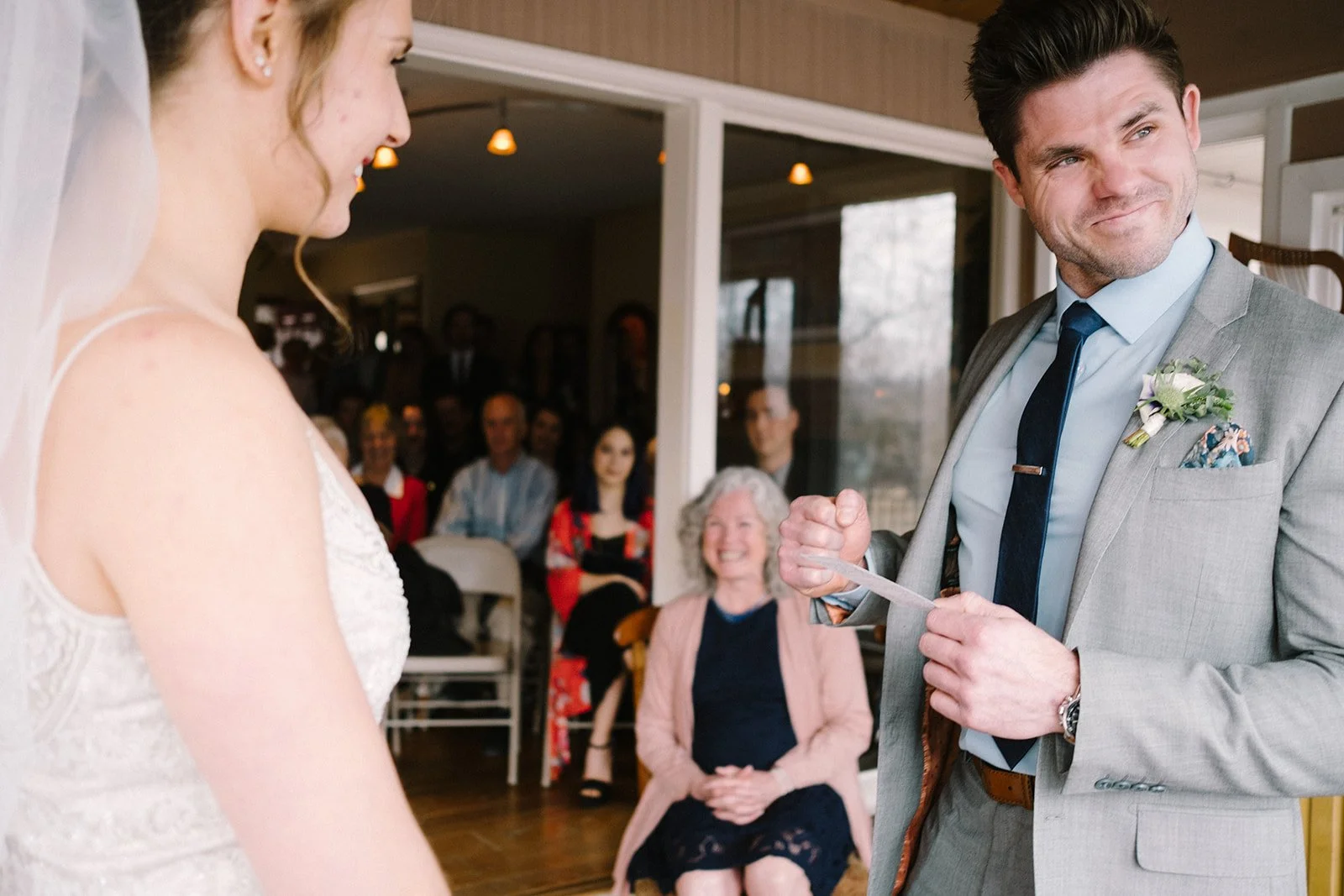 A groom in a gray suit and a bride in a white wedding dress during a wedding ceremony. The groom is holding a piece of paper, smiling, as he looks at the bride. The bride is smiling back. Guests are sitting in the background, watching the ceremony wi