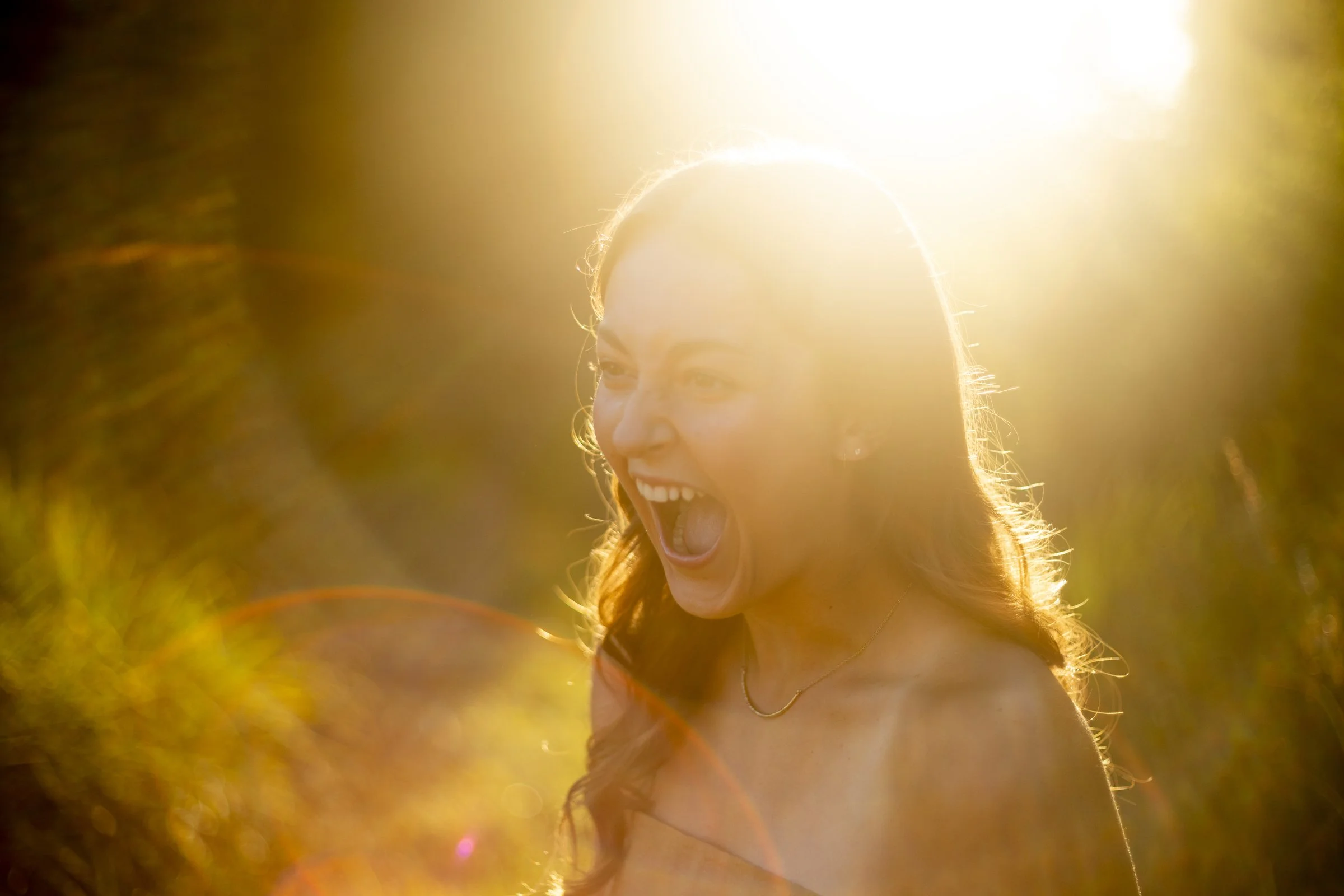 A young woman with long brown hair smiling and laughing outside during sunset, illuminated by warm sunlight.