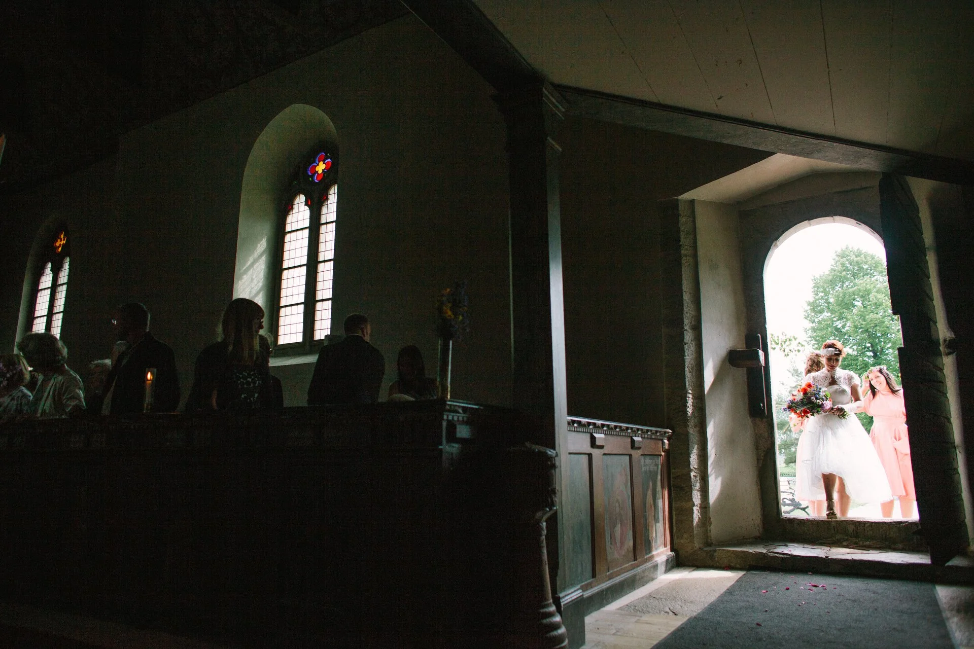 Inside a dimly lit church, guests sit on pews while a bride in a white wedding dress and veil, holding a bouquet, stands near the entrance with another woman in a pink dress, with the outside visible through an open door and a large tree in the backg