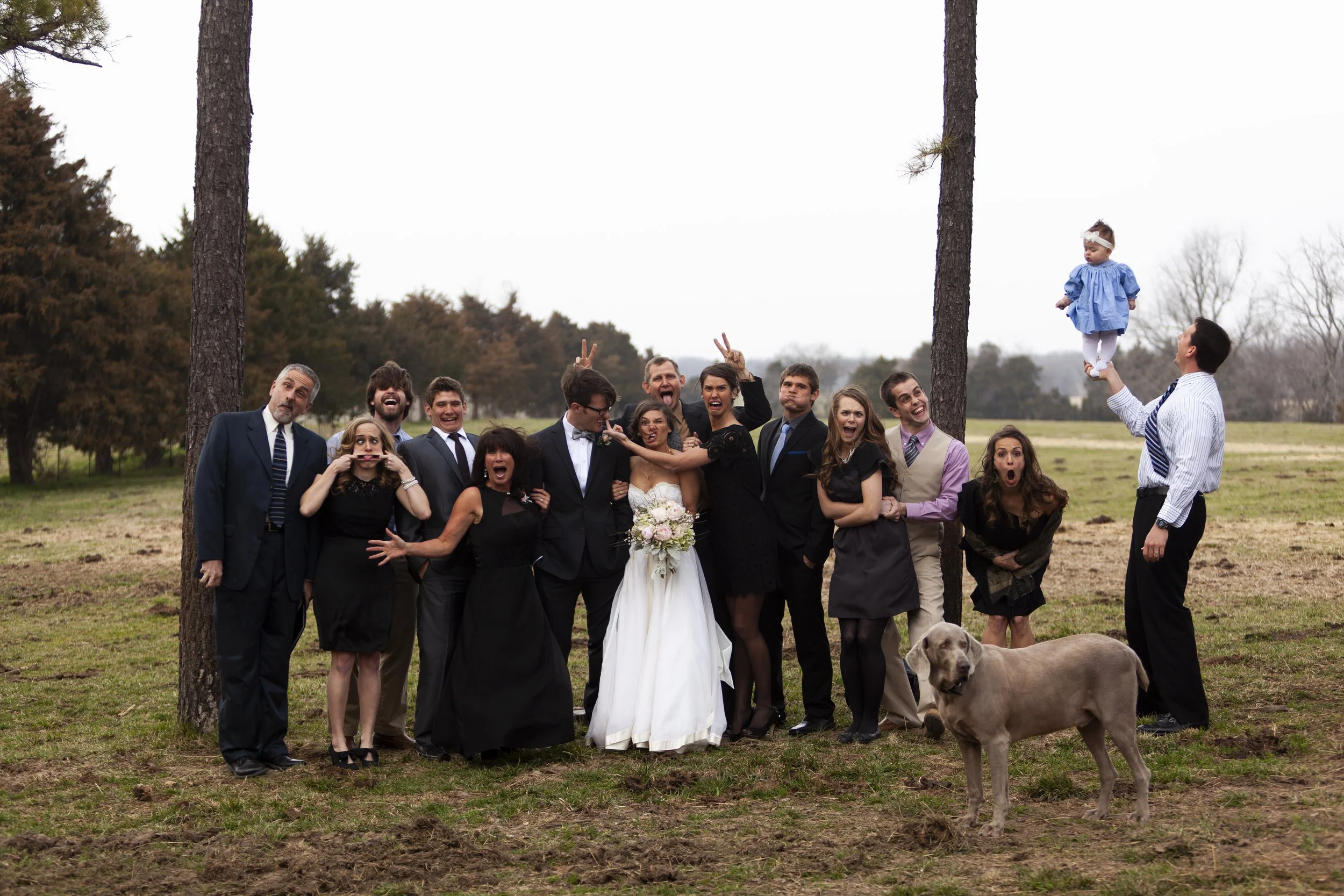 A group of people at a wedding outdoor gathering, with a bride and groom in the center, some making funny faces or gestures, with trees and an overcast sky in the background. A man is lifting a young girl in a blue dress into the air, and a dog is ly