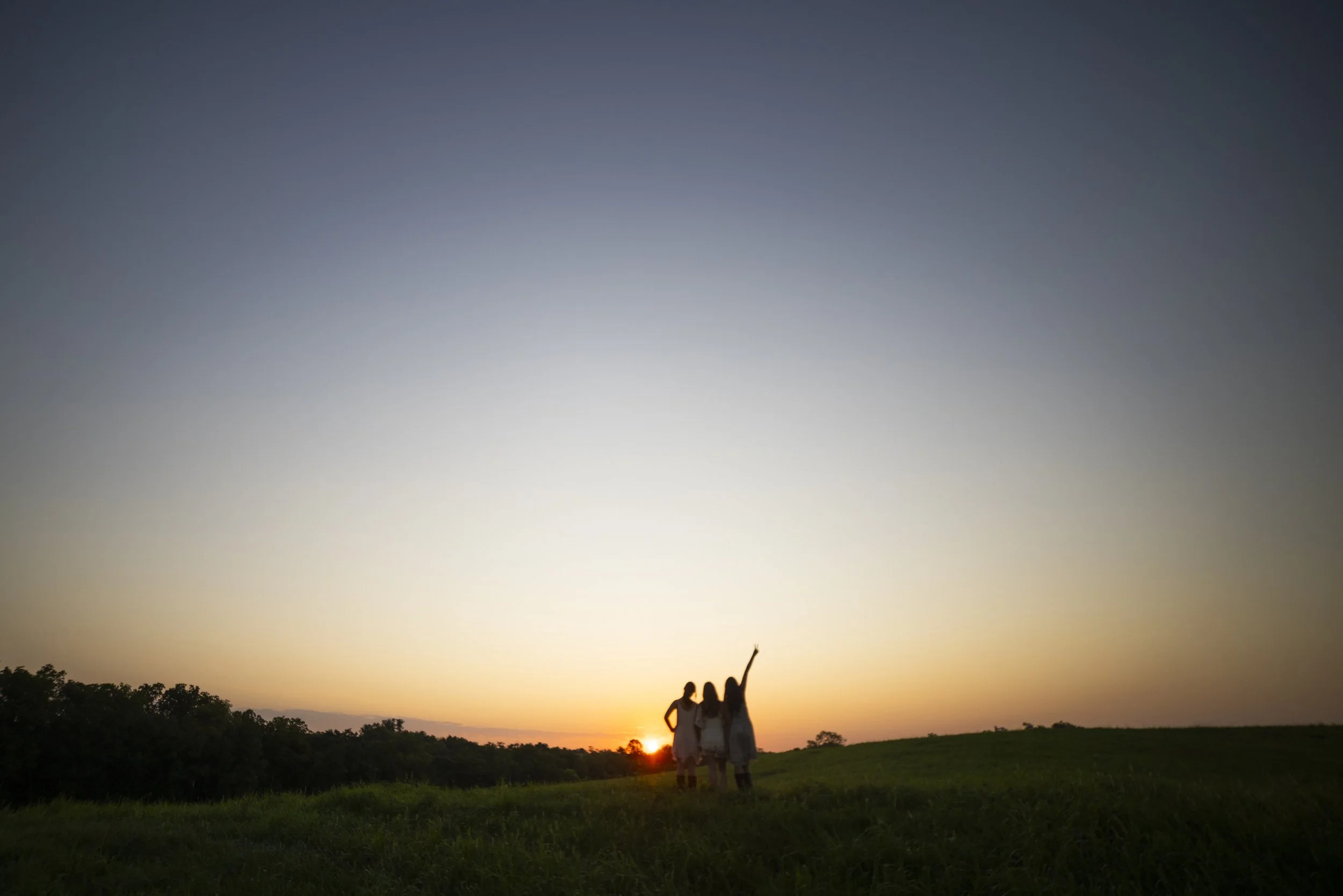 Three women walking together on a grassy hill at sunset, with the sun setting behind them and a clear sky above.