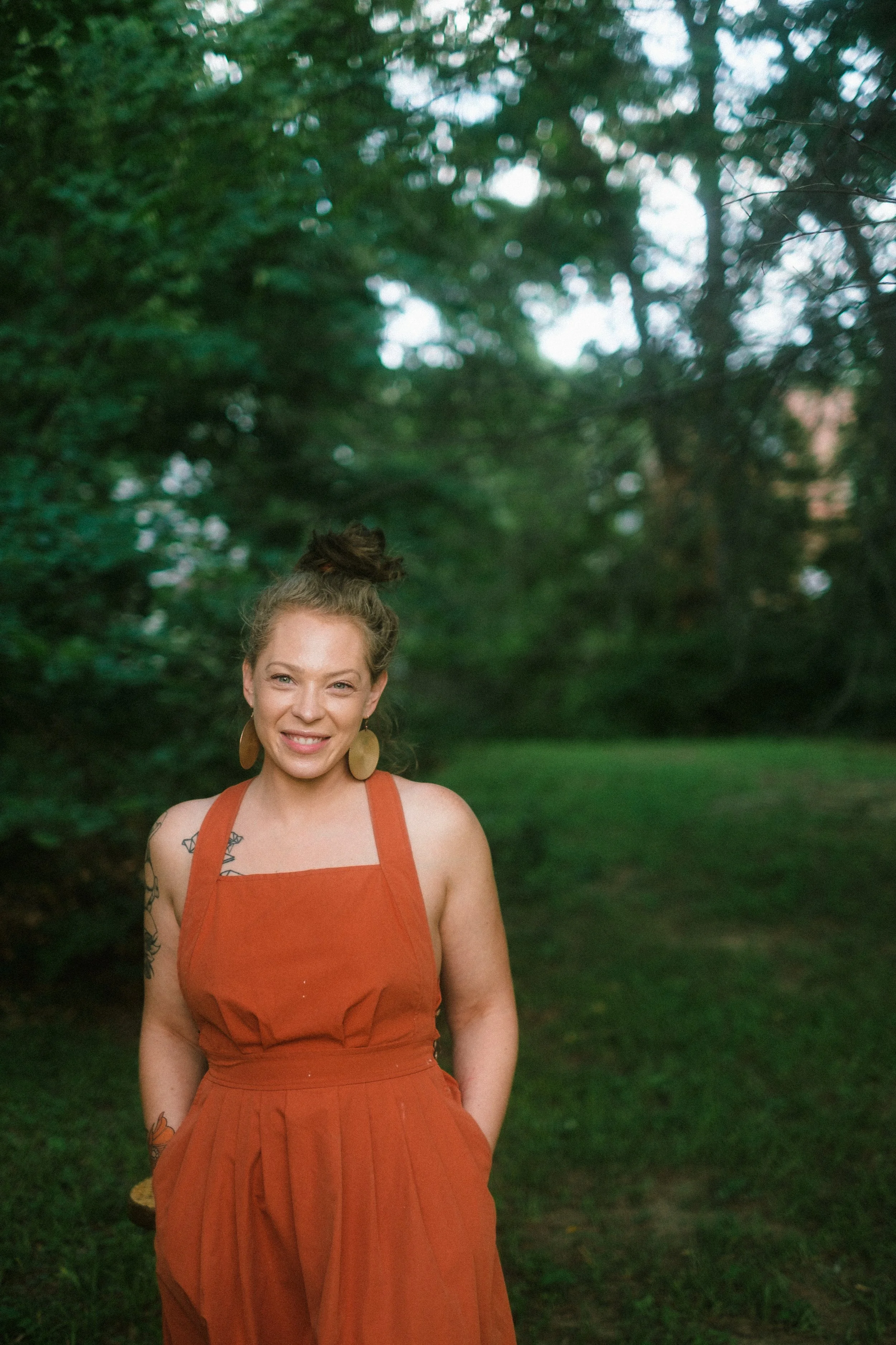 A woman standing outdoors in a park with green trees in the background. She has light skin, brown hair styled in a bun, and is wearing a sleeveless orange dress with large earrings.