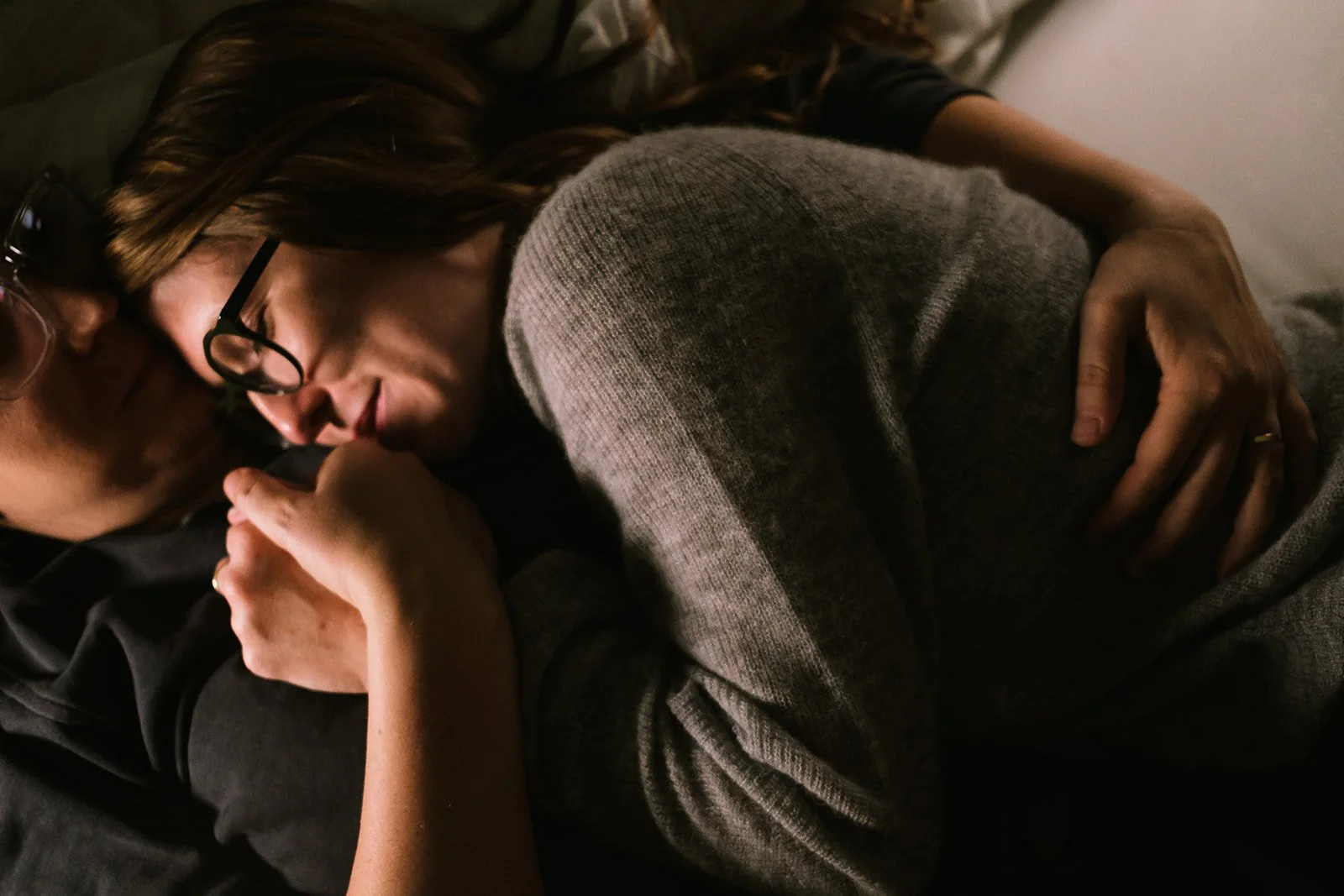 A woman and a man are lying on a bed, closely embracing. The woman has long hair, glasses, and is resting her head on the man's shoulder while holding a phone. The man has glasses and is wearing a dark shirt. The lighting is soft and warm.