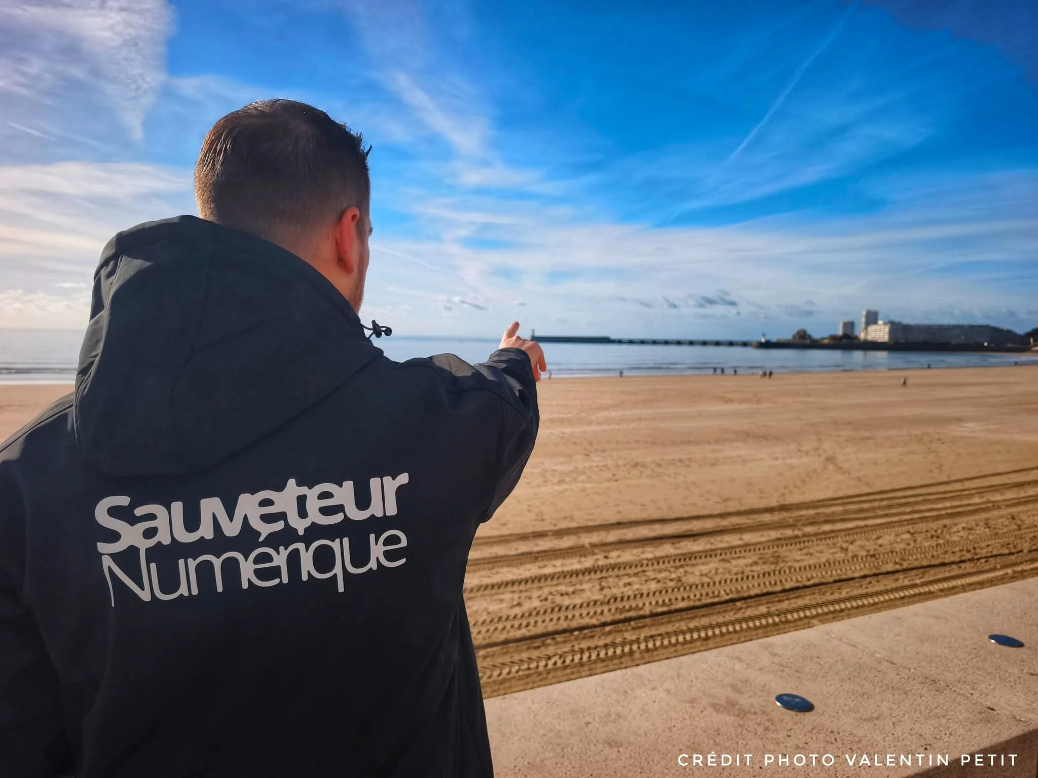 Un homme vu de dos portant une veste noire avec l'inscription « Sauveur Numérique » qui pointe du doigt vers la mer sur une plage ensoleillée avec un ciel bleu et quelques nuages, horizon avec des bâtiments.