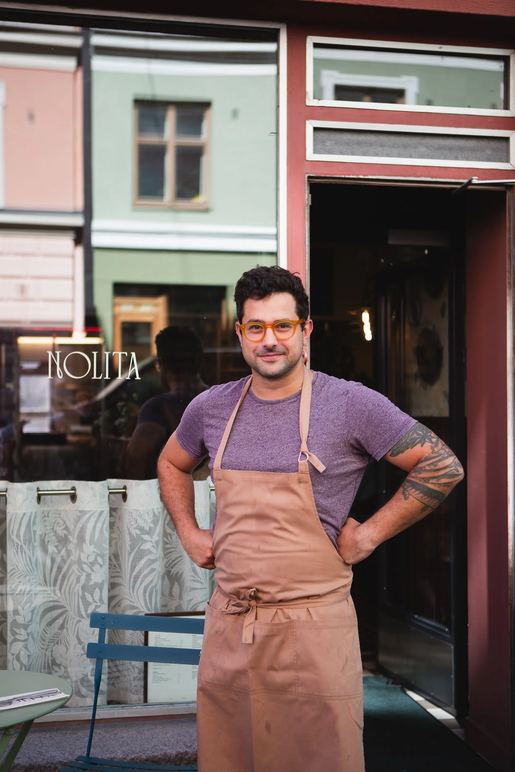 A man with dark curly hair and glasses, wearing a purple t-shirt and a tan apron, standing outside a restaurant with one hand on his hip, smiling, in front of a window with the word 'NOLITA' on it.