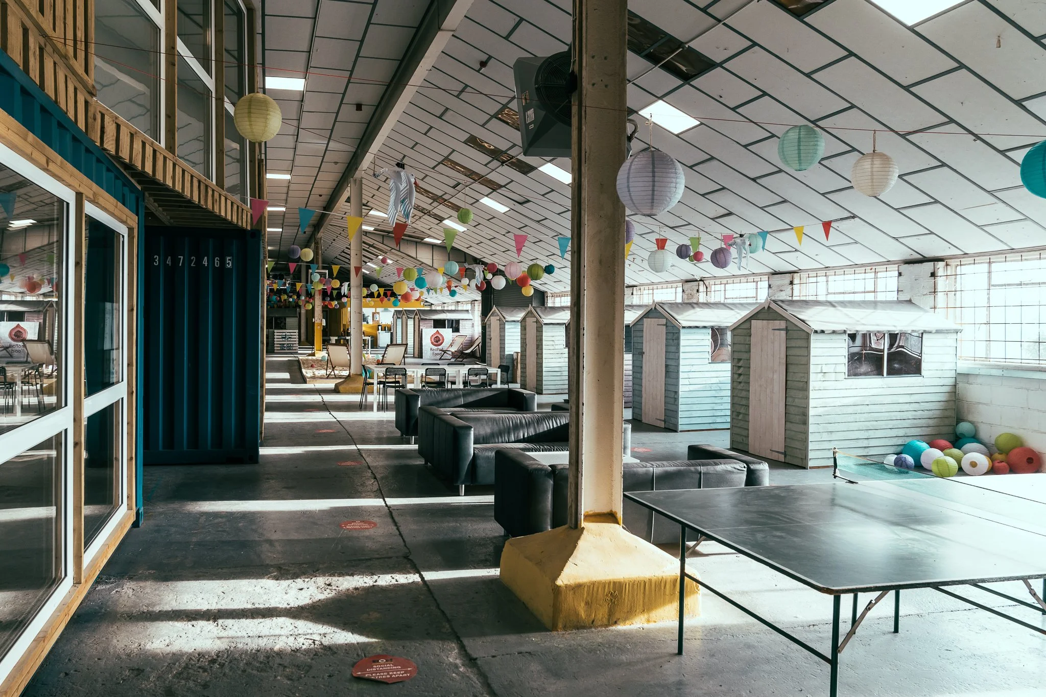 Indoor recreational space decorated with colorful paper lanterns, buntings, and small houses, featuring black leather couches, a ping pong table, and a ball pit, illuminated by natural light.