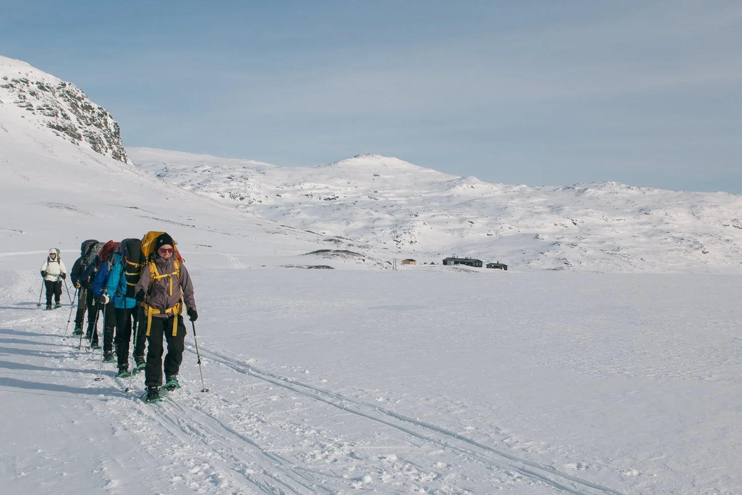 A group of people in winter gear cross-country skiing on snow in a mountain landscape with snow-covered hills, a few distant buildings, and a partly cloudy sky.