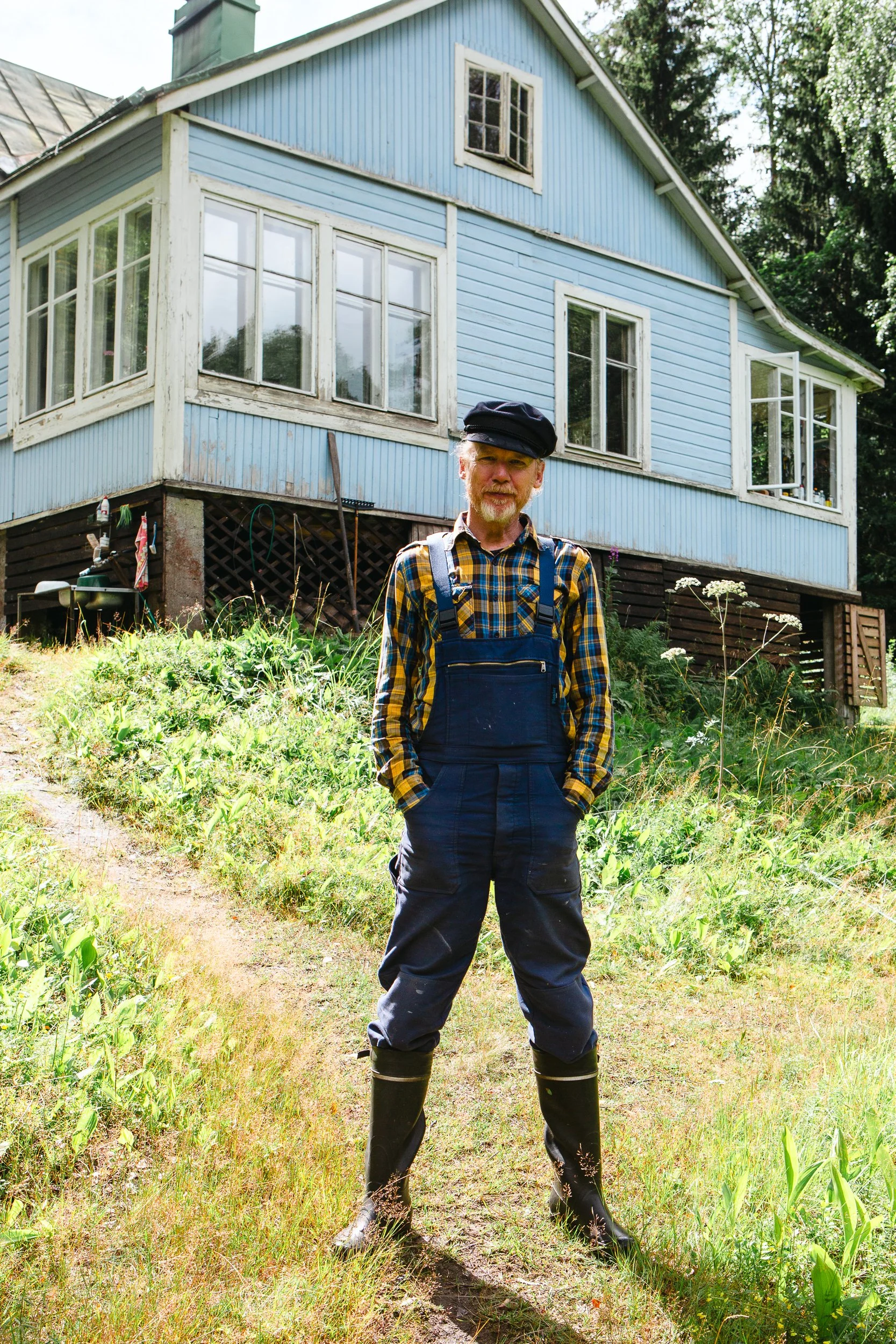 A man standing outdoors in front of a light blue house with white trim, wearing a plaid shirt, overalls, and rubber boots.