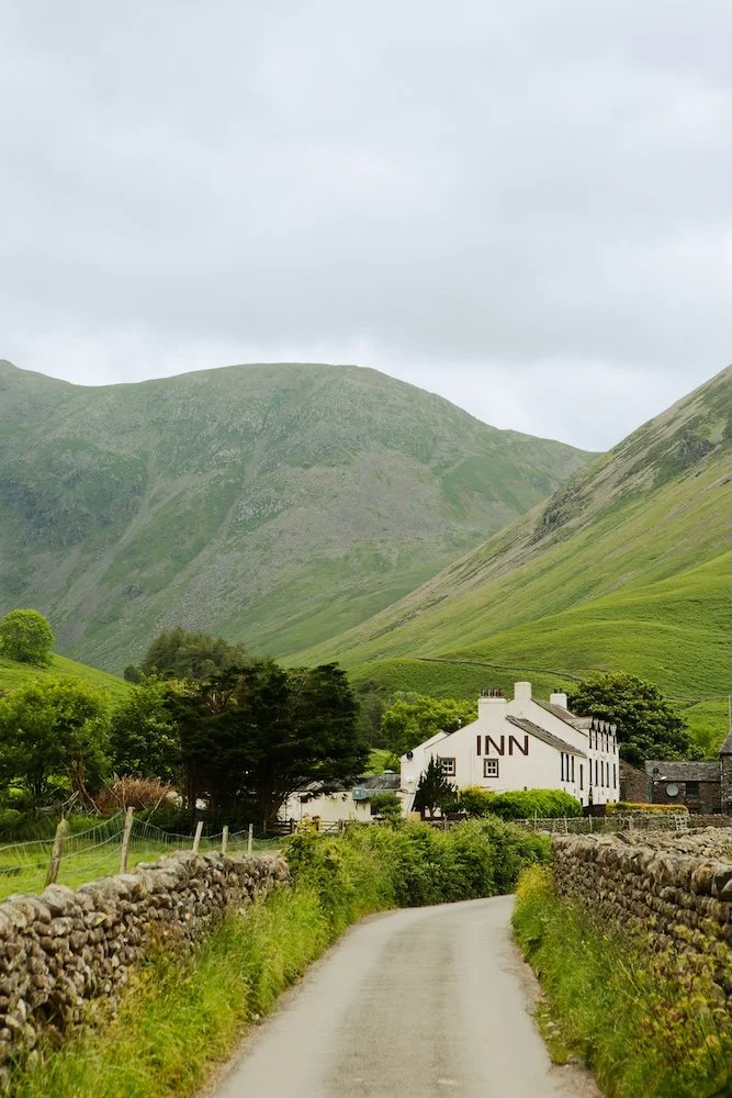 A rural scene with a narrow country road leading to a white inn building at the base of green hills under a cloudy sky.