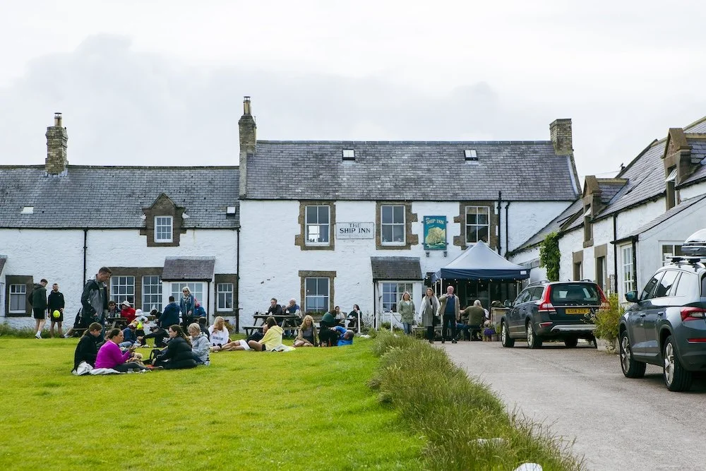 People gathered outside the Ship Inn pub, sitting on the grass and socializing, with cars parked on the driveway, and the pub building in the background.