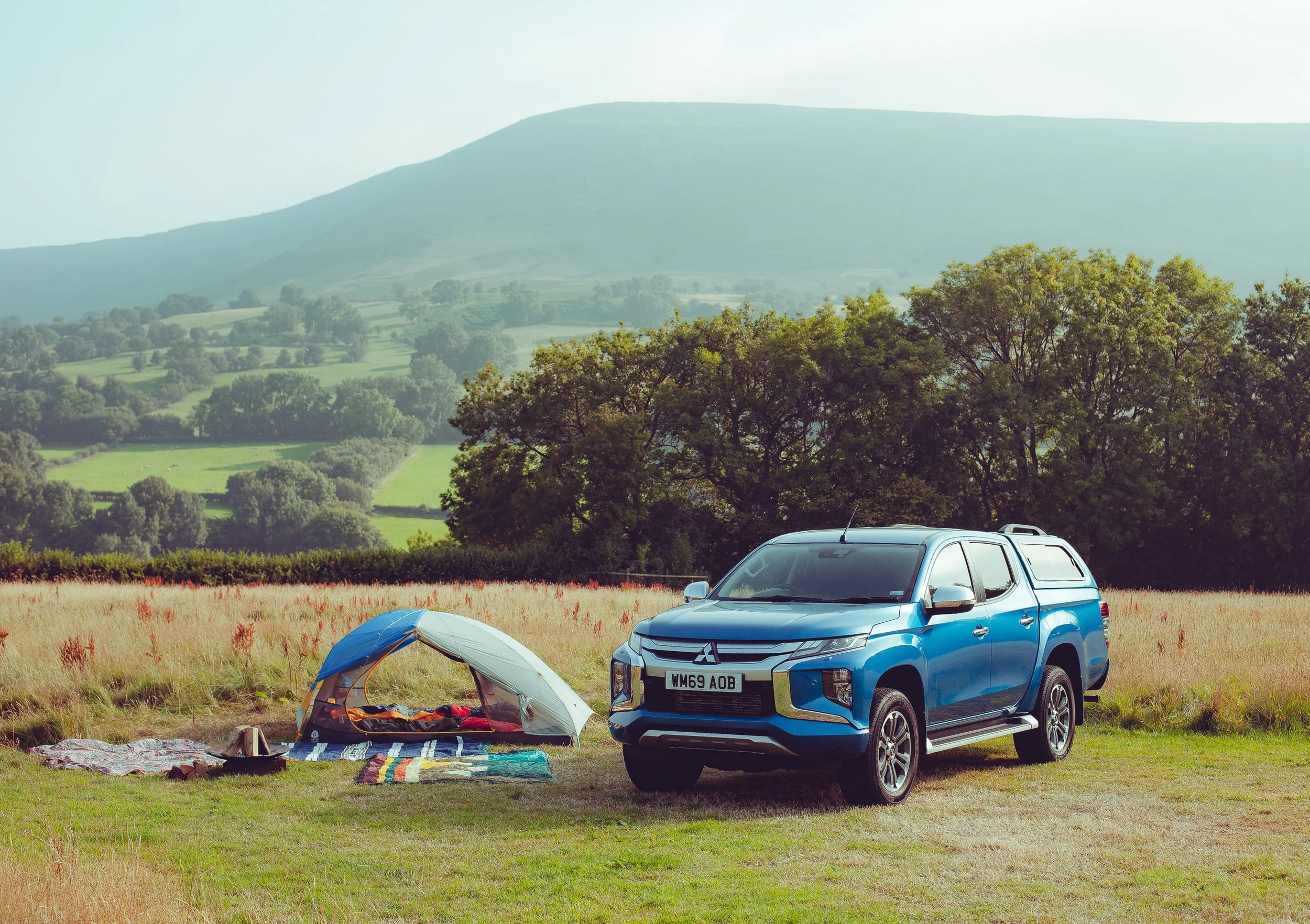 Blue pickup truck parked on a grassy field near a small camping tent with blankets and belongings, against a backdrop of rolling hills, trees, and distant mountains.