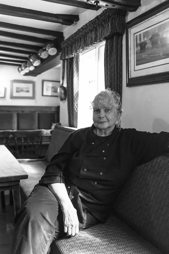 An elderly woman with curly hair sitting on a patterned couch in a cozy, traditional room with framed pictures, curtains, and a window.