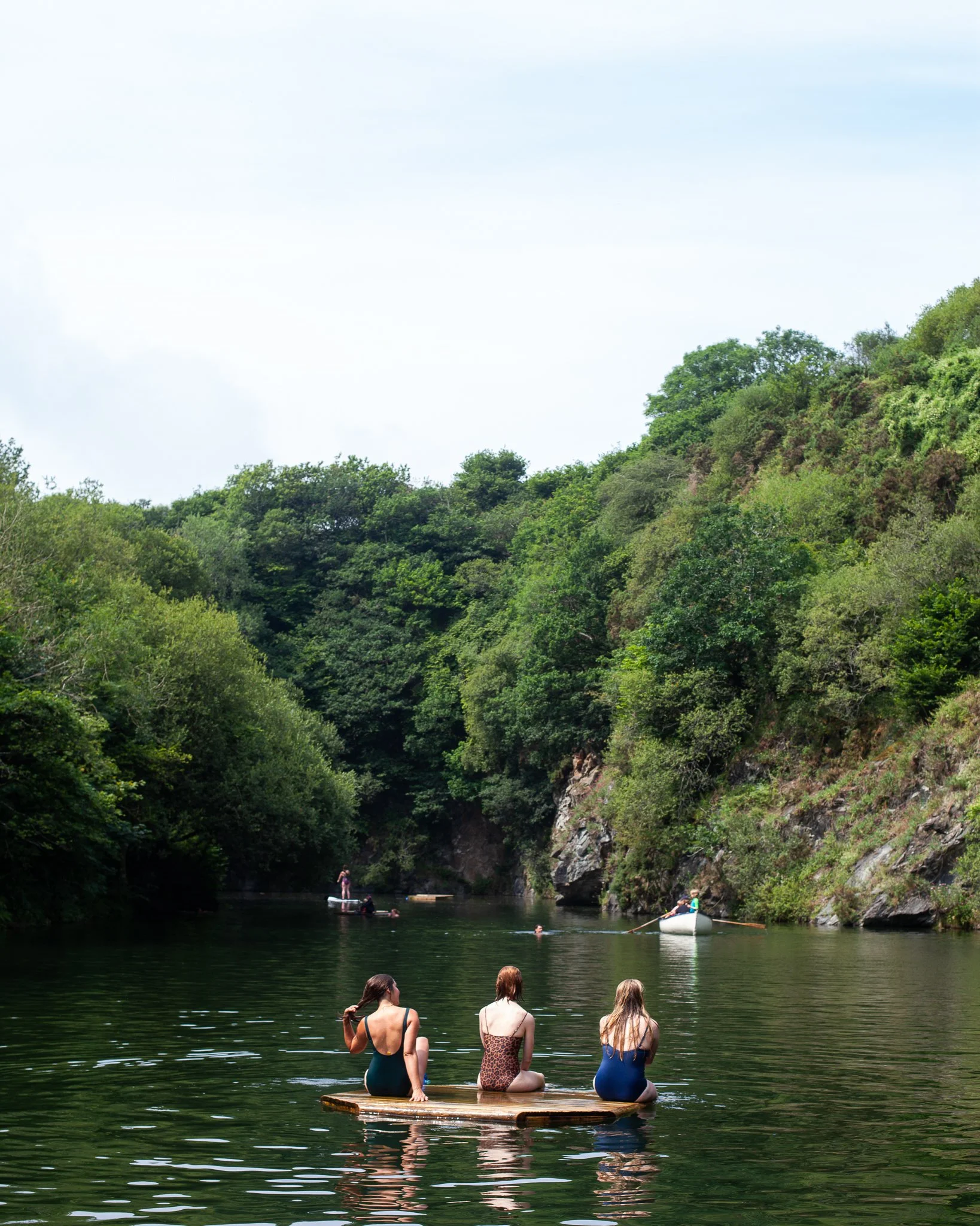 Three women sit on a wooden raft in a river surrounded by lush green trees and cliffs; others are in boats and swimming in the river.