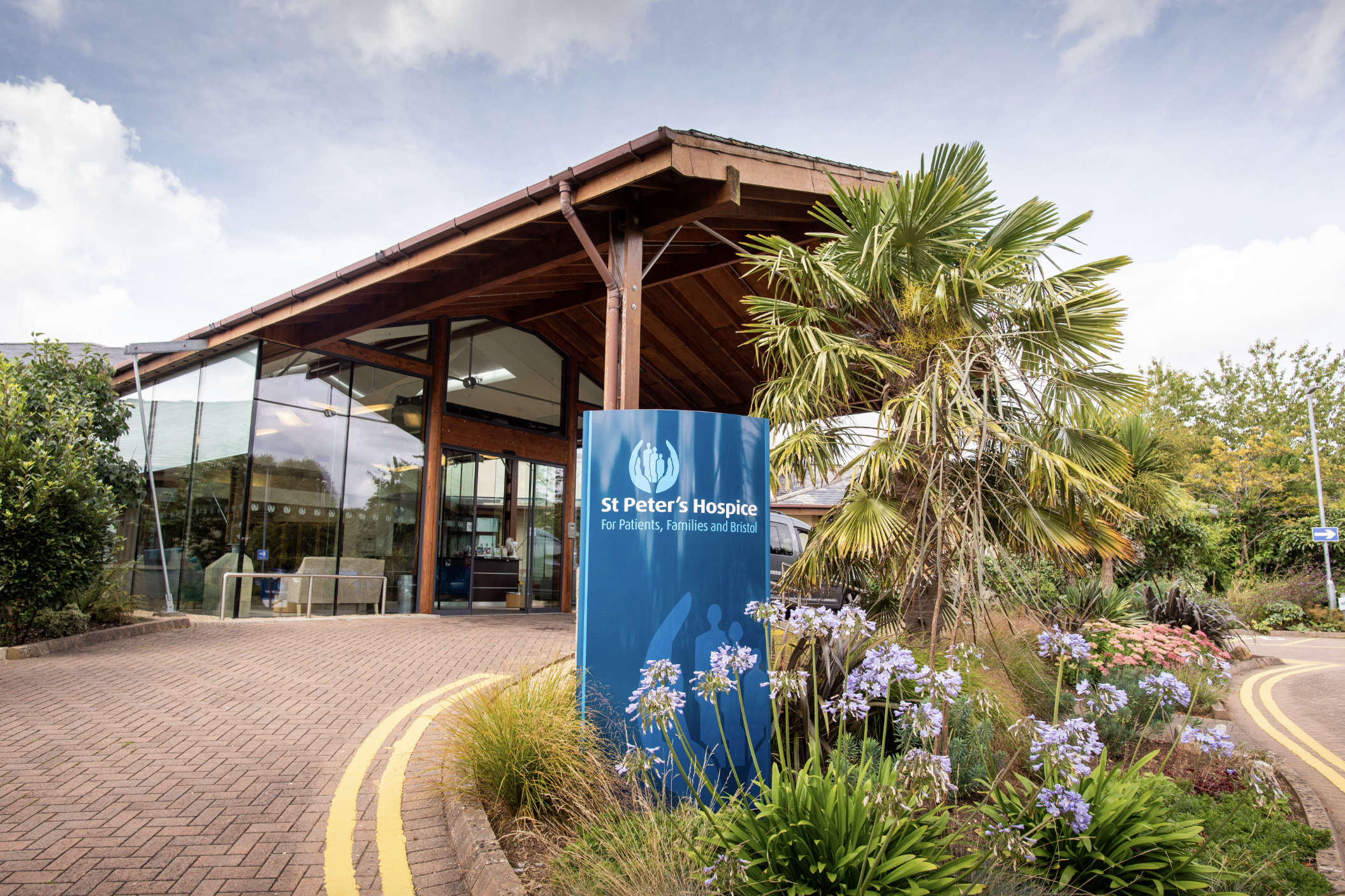Entrance of St Peter's Hospice facility with a blue sign and surrounding landscaped garden with purple flowers and a palm tree.