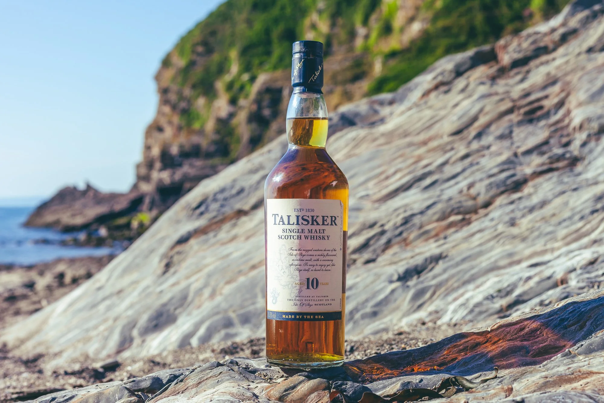 A bottle of Talisker single malt Scotch whisky placed on a rock by the seaside with a rocky cliff and ocean in the background.