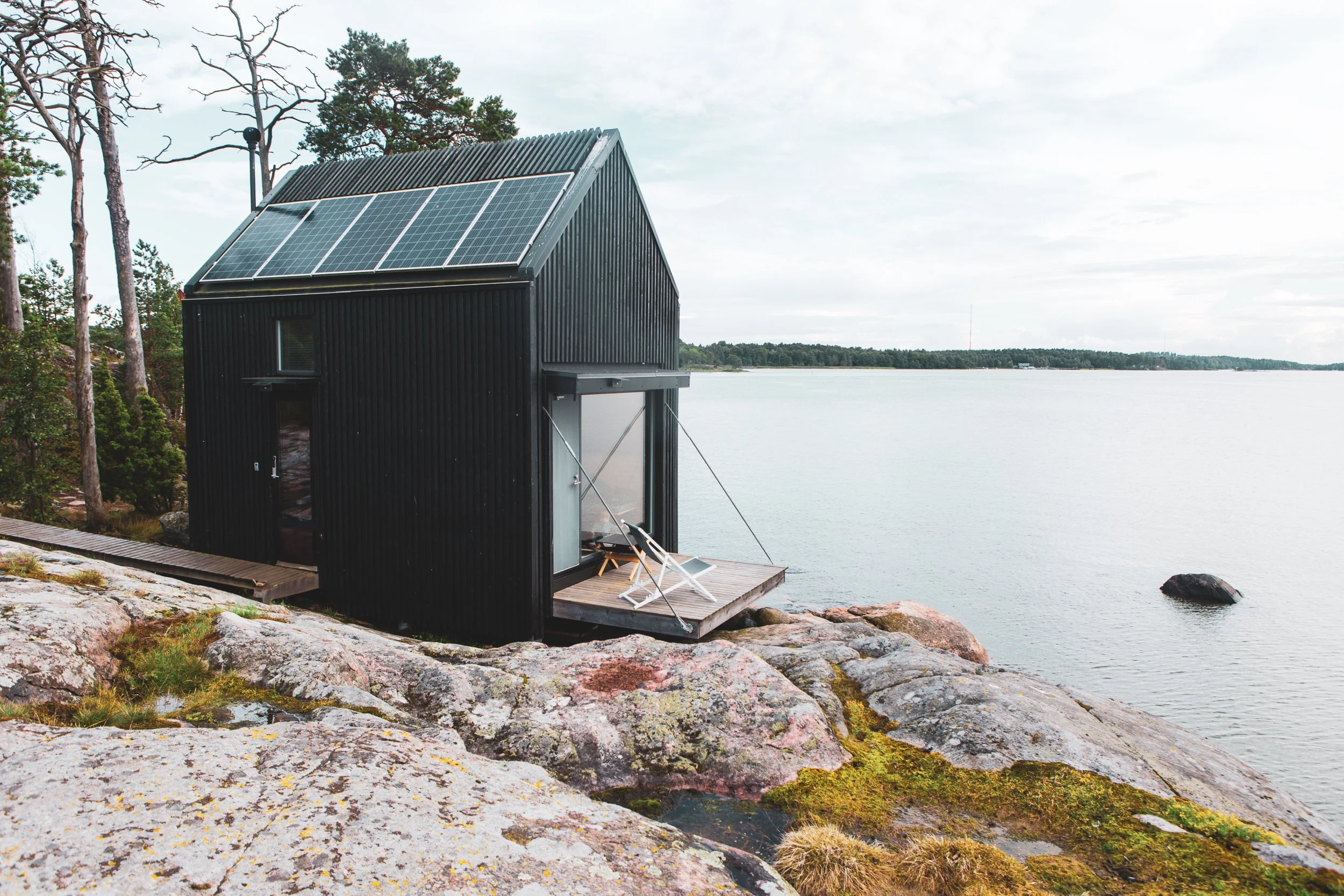 A black tiny house with solar panels on the roof, located on a rocky shoreline next to a large body of water, with a deck that has a lounge chair, surrounded by trees.