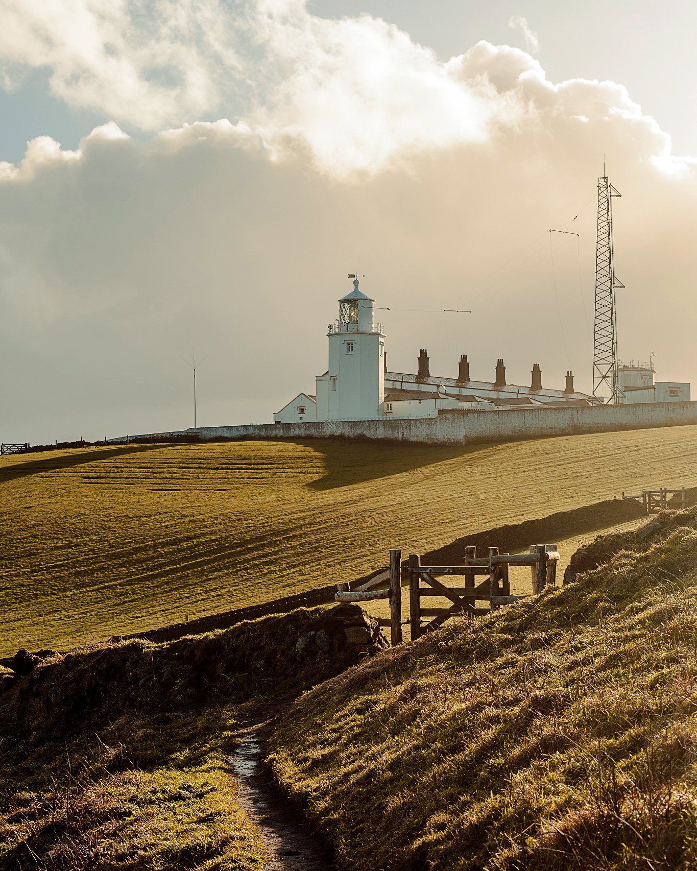 A lighthouse on a hill with green grass, a dirt path and a wooden fence in the foreground, and a partly cloudy sky in the background.