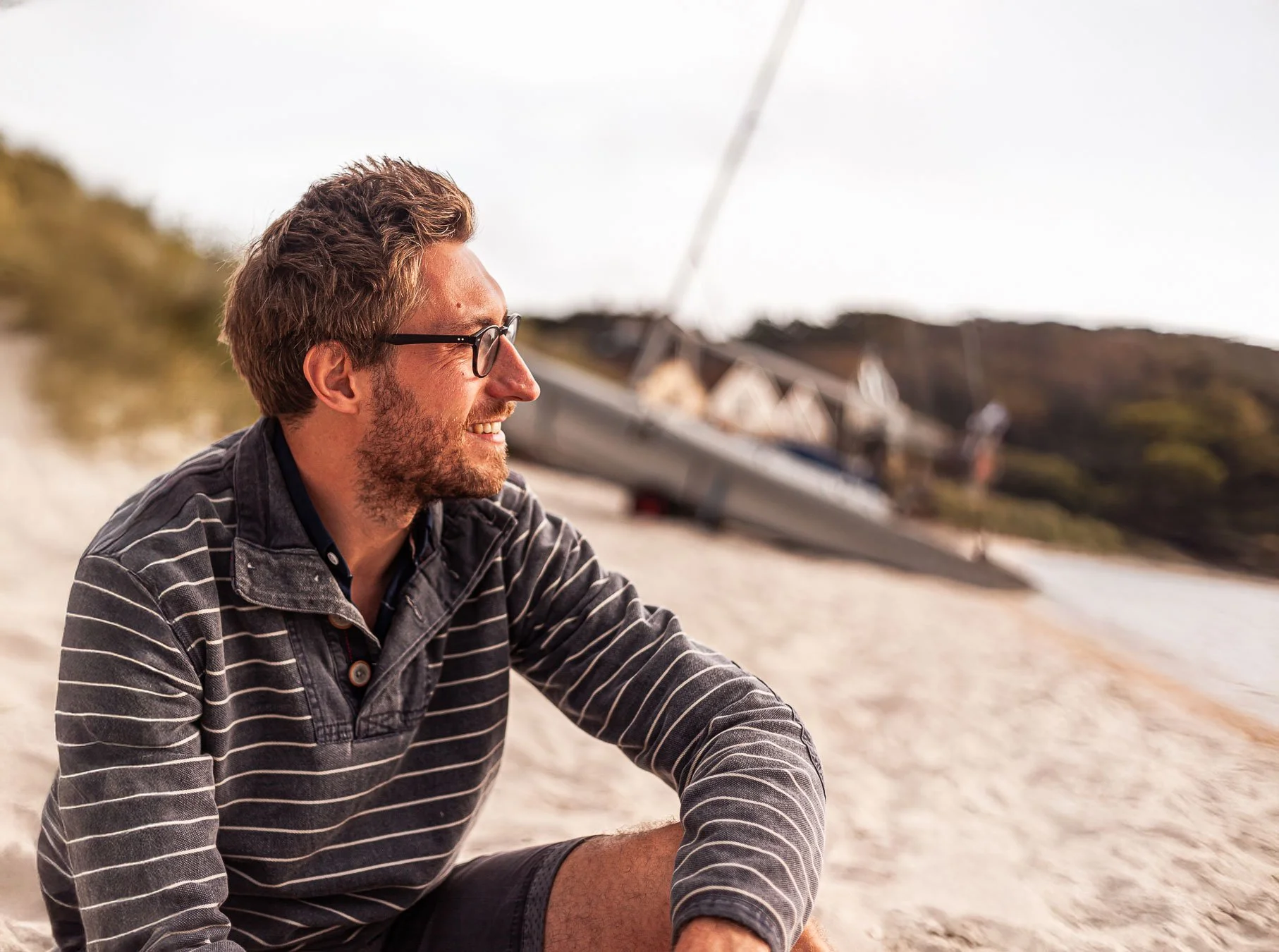 A man with glasses and a beard sitting on a sandy beach, smiling and looking to the side, with sailboats and a hillside in the background.