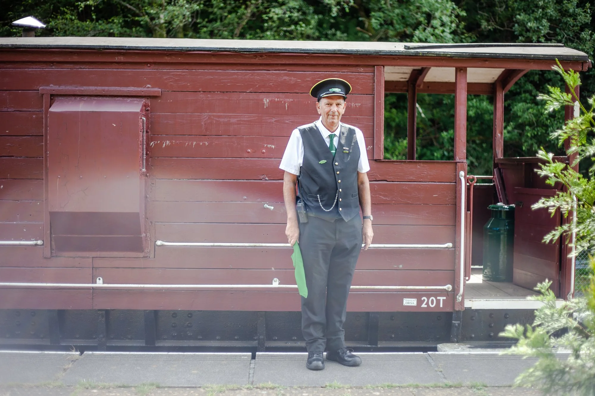 A man dressed in a vintage train conductor uniform standing in front of a red wooden train car, holding a green cloth.