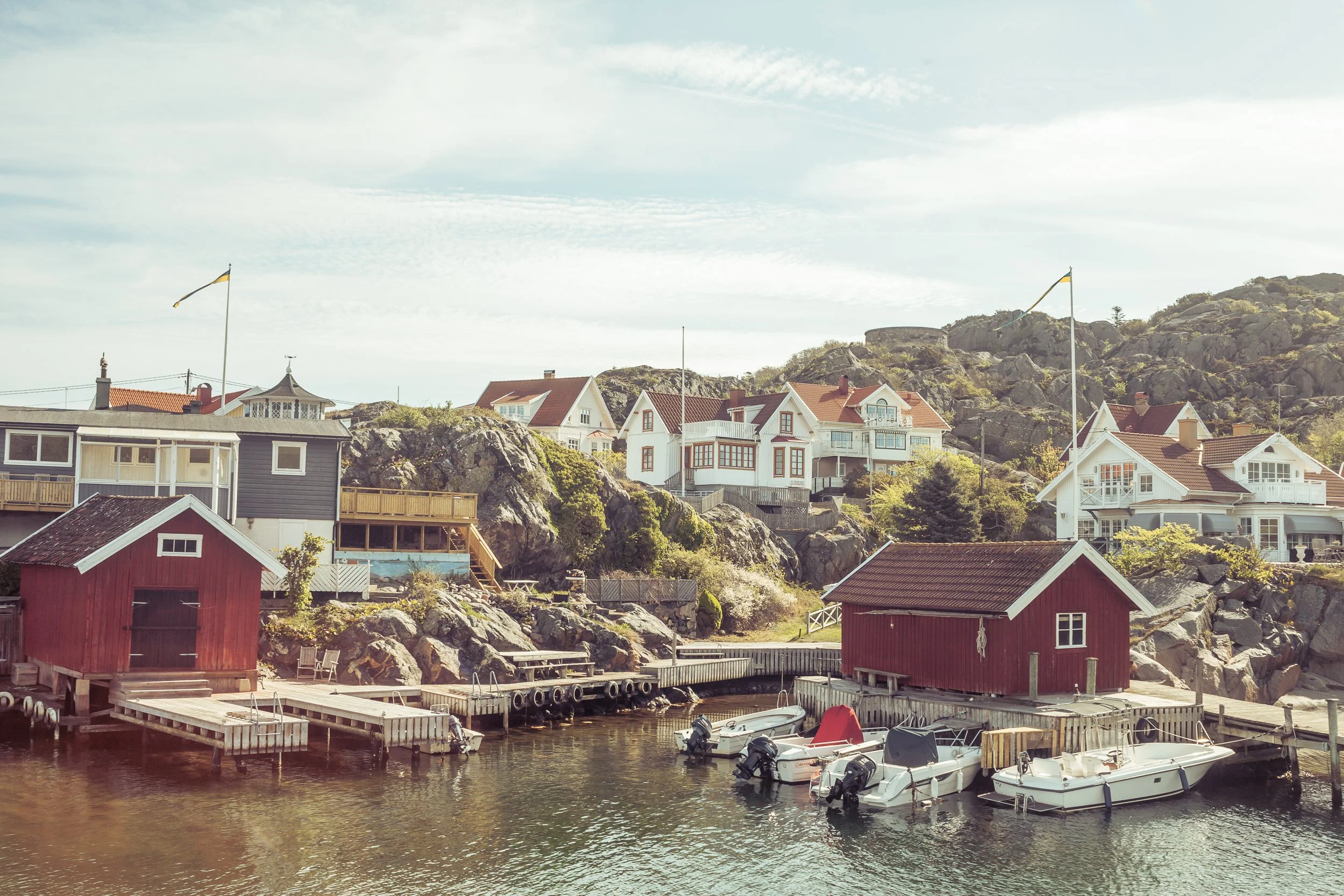 Coastal village with colorful houses, a small marina with boats, and rocky hills in the background under a partly cloudy sky.
