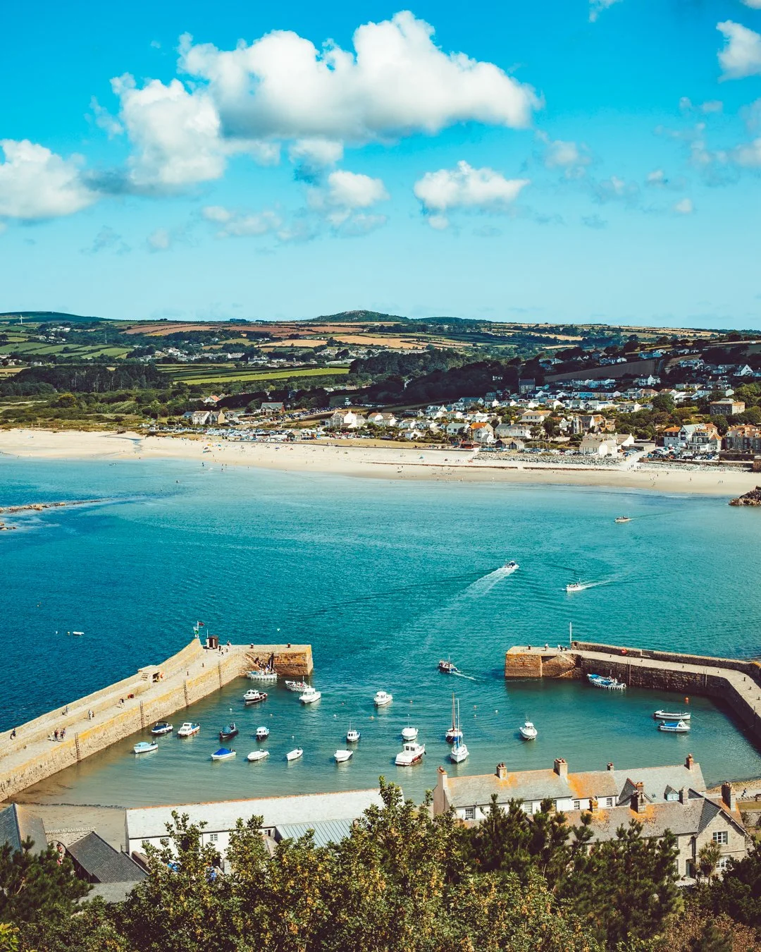 A coastal town with a harbor filled with boats, sandy beach, green hills, and houses along the shoreline, under a blue sky with scattered clouds.