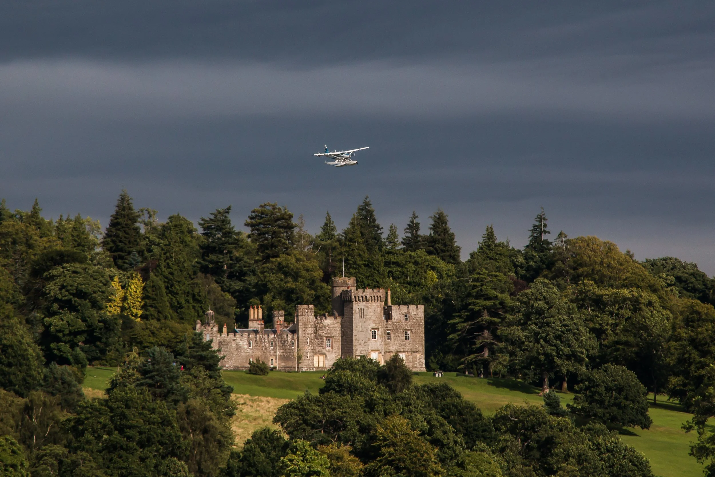 A castle situated on a hill surrounded by dense trees with a small airplane flying overhead against a dark, cloudy sky.