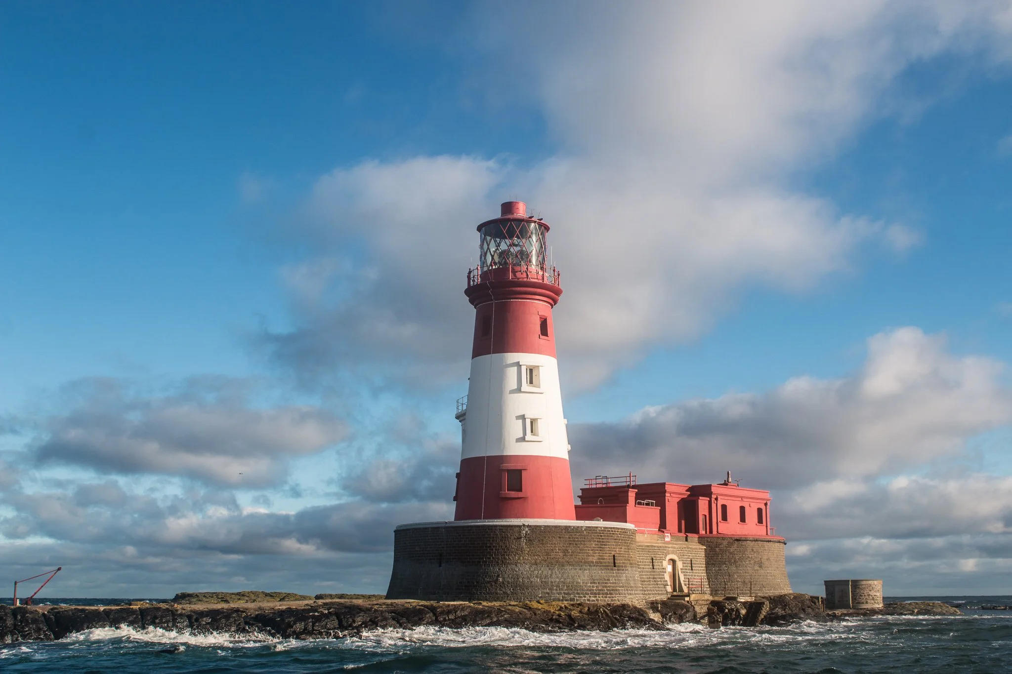 A red and white striped lighthouse on a rocky island with a building attached, under a partly cloudy sky.