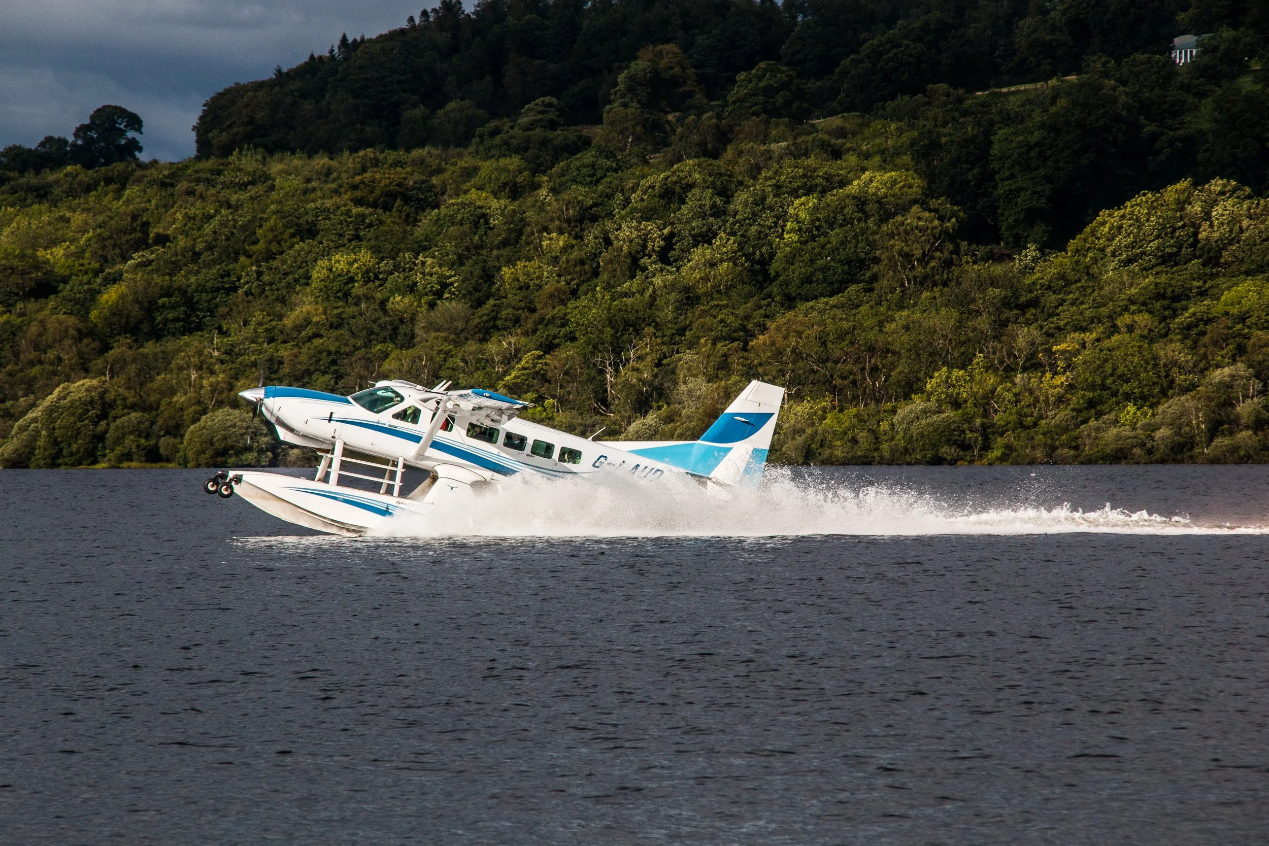 A seaplane skimming the water's surface during takeoff or landing with a lush forested hillside in the background.