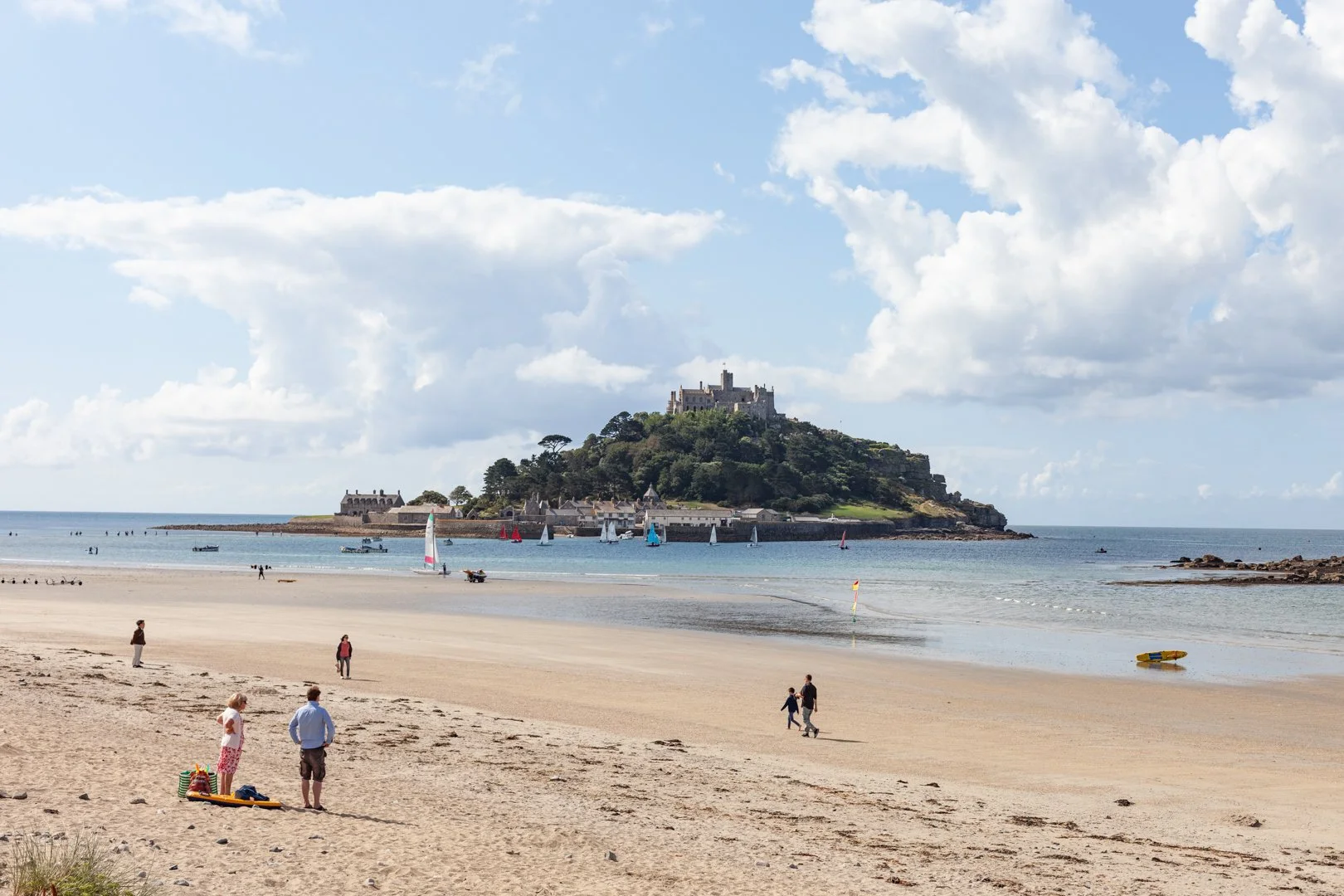 Scenic view of a beach with people walking and relaxing. In the background, St. Michael's Mount castle on a hill, with sailboats on the water and a partly cloudy sky.