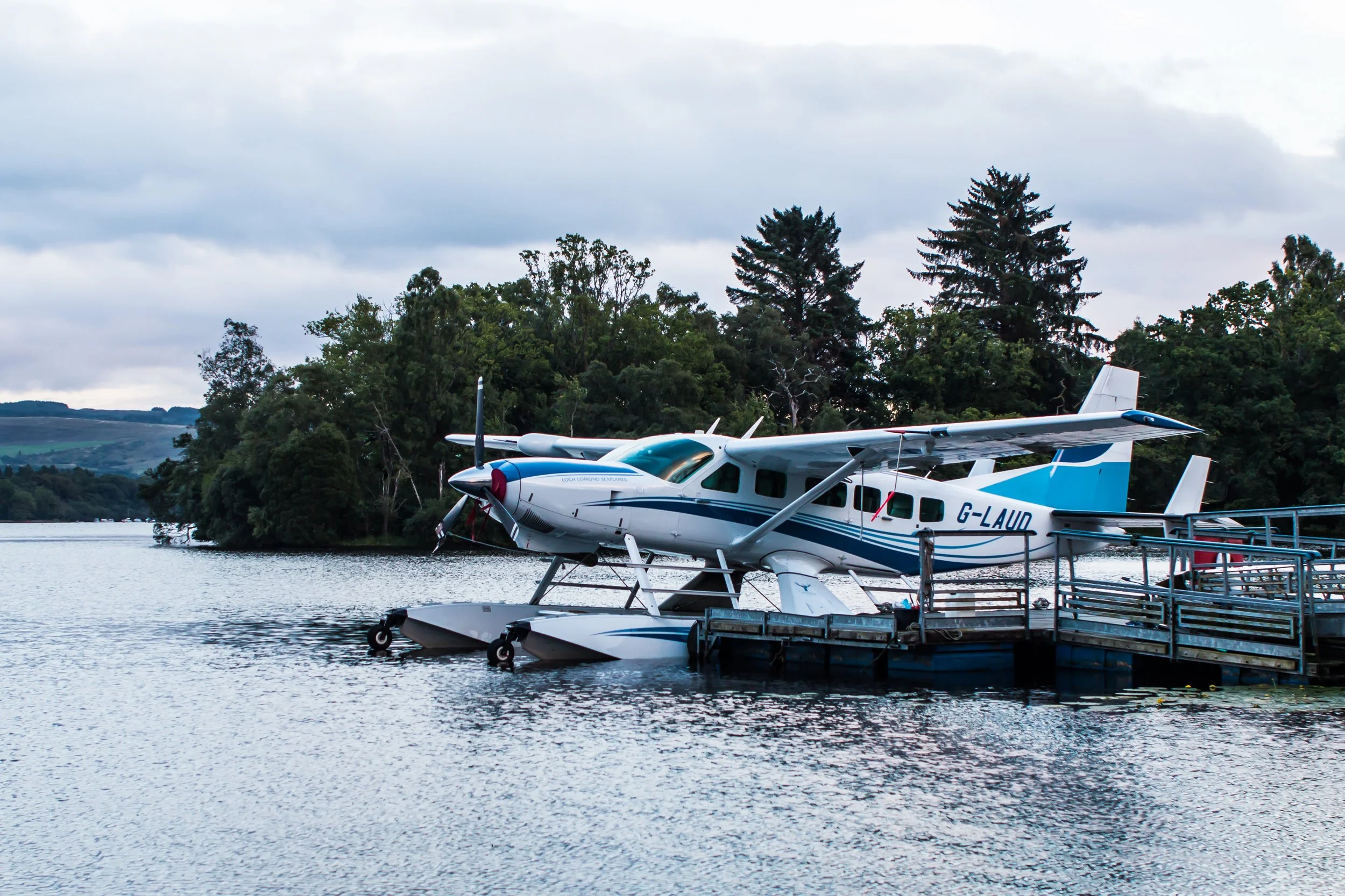 A seaplane docked at a wooden pier on a lake, surrounded by trees and cloudy sky.