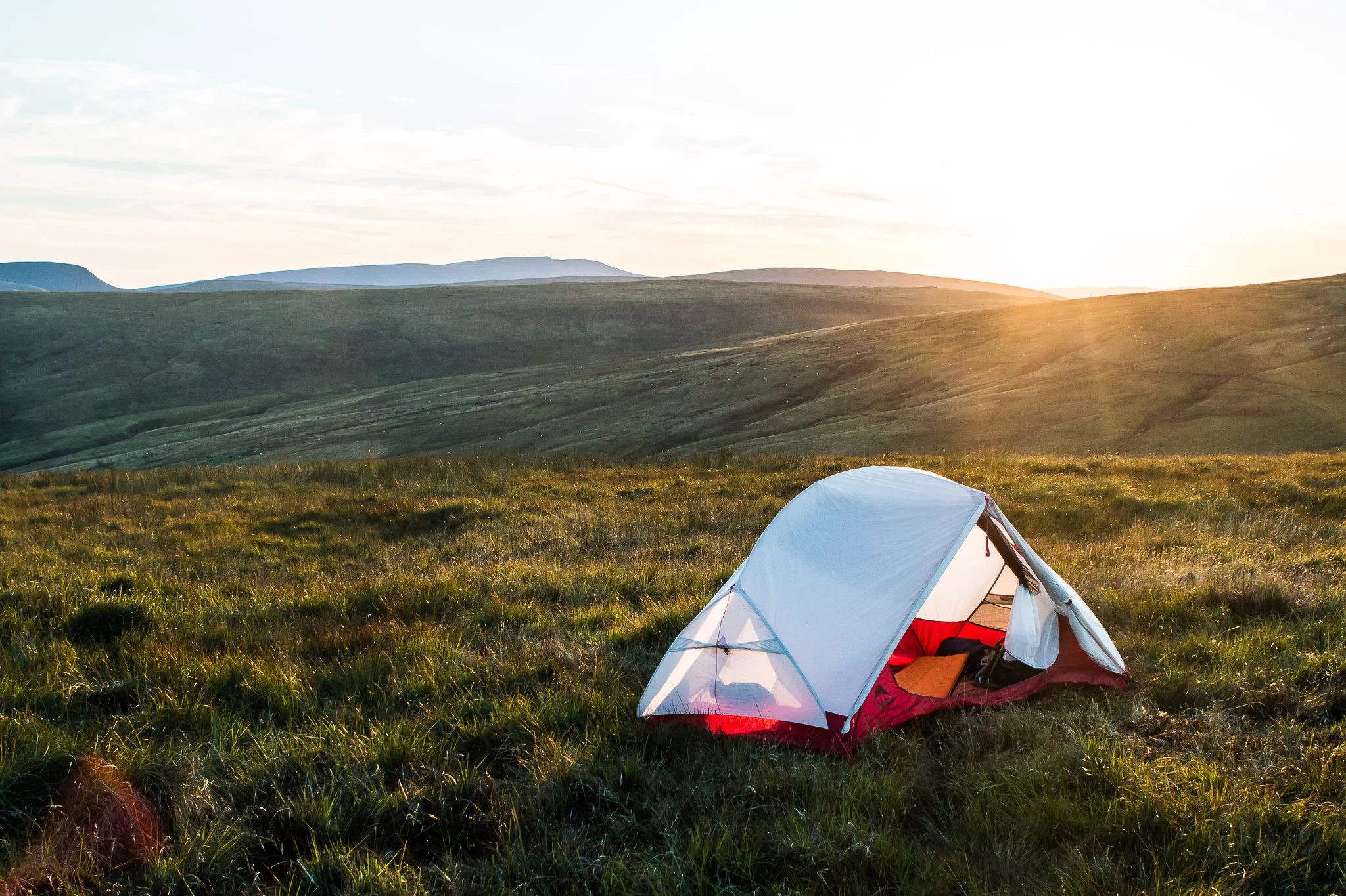 A white and red camping tent on a grassy field at sunset with rolling hills in the background.