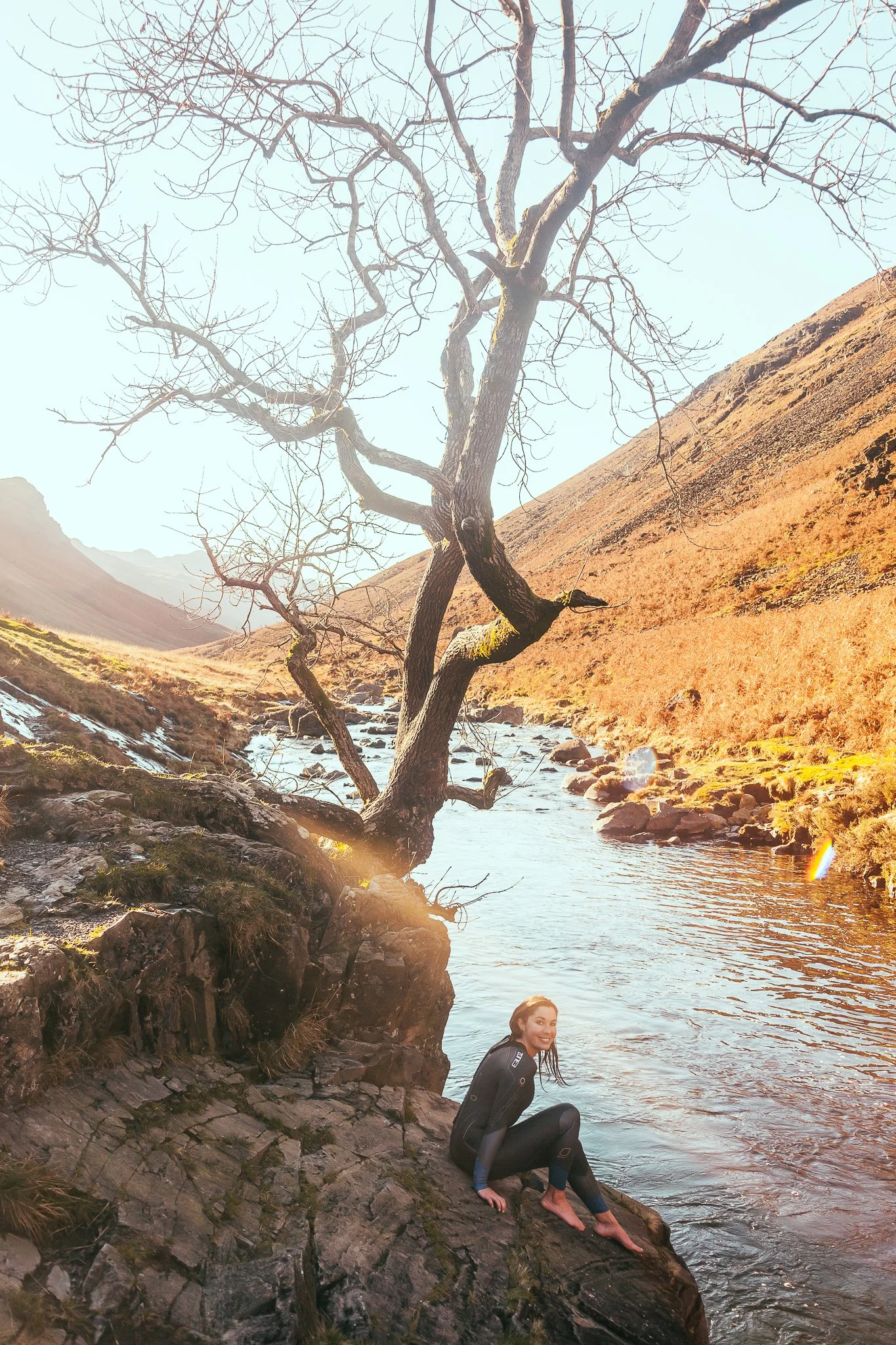 A woman sitting on a rock by a river in a mountainous area with a leafless tree and a clear sky in the background.