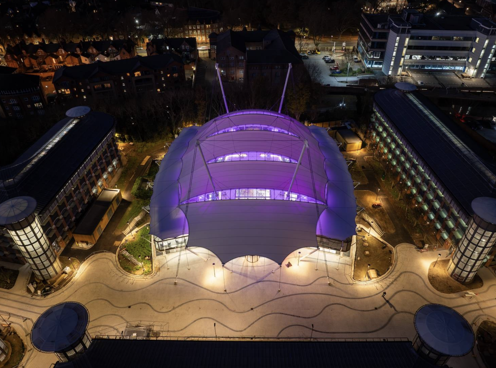 Aerial nighttime view of a modern building with a large, brightly lit, curving roof structure illuminated with purple lights, surrounded by other buildings and streets.