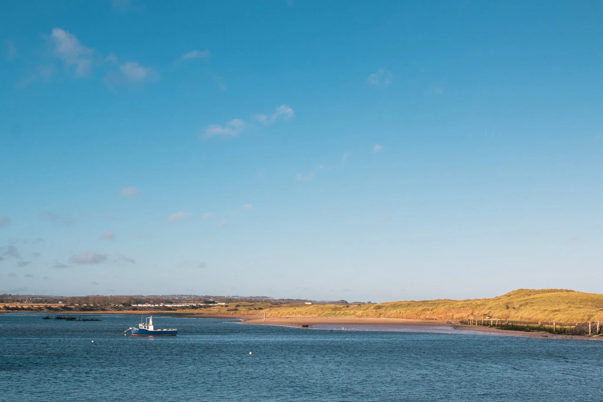 A calm body of water with a boat near the shore, grassy land in the background, and a blue sky with a few clouds.