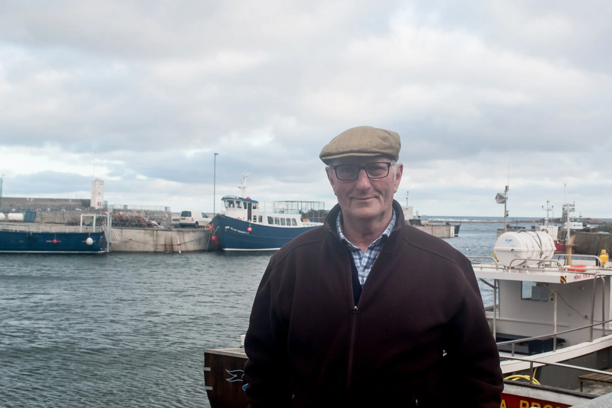 An older man wearing a flat cap, glasses, and a dark jacket standing near a harbor with boats docked in the background under a cloudy sky.