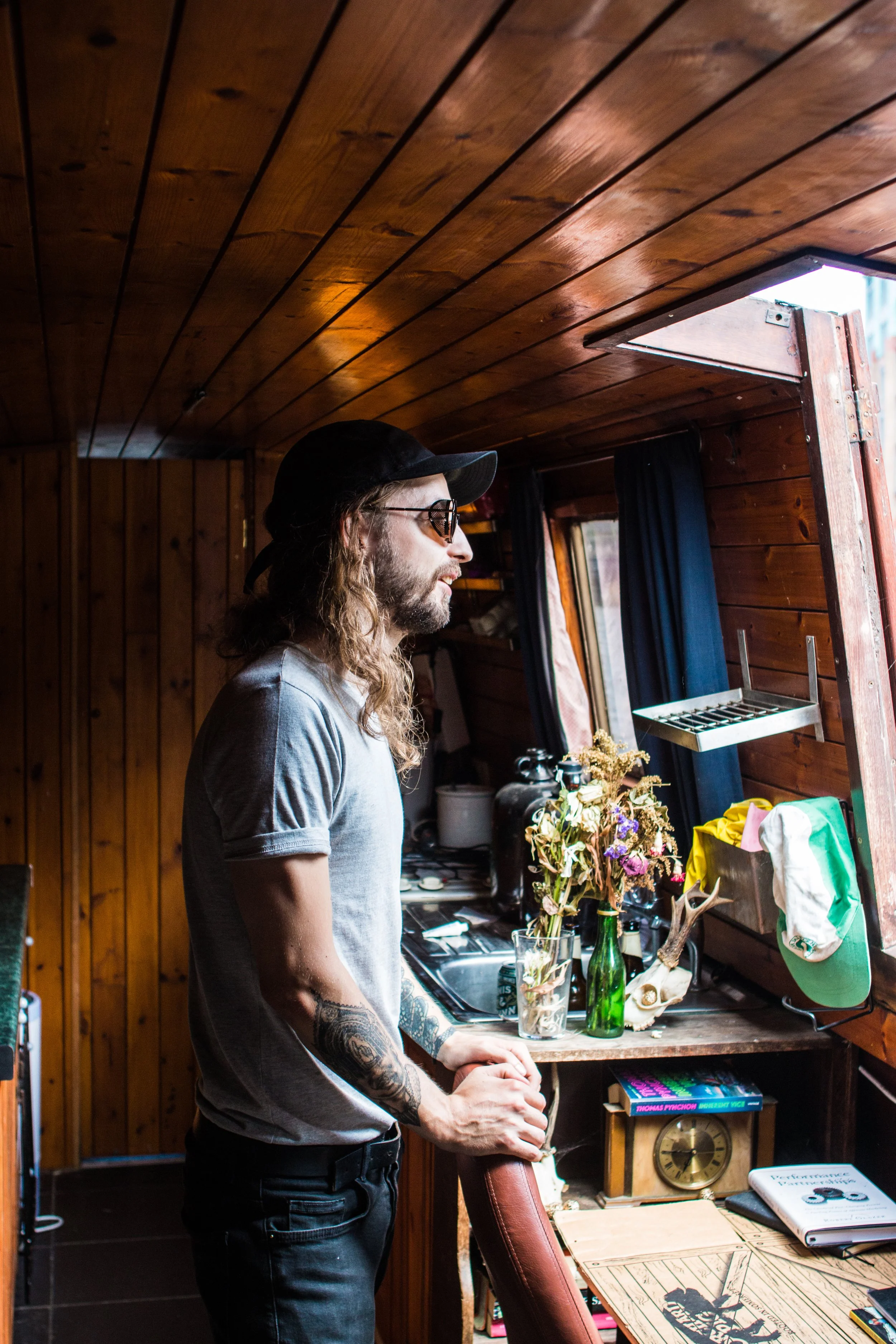 A man with long hair, beard, sunglasses, and tattoos is standing in a wood-paneled room near a window, looking outside.