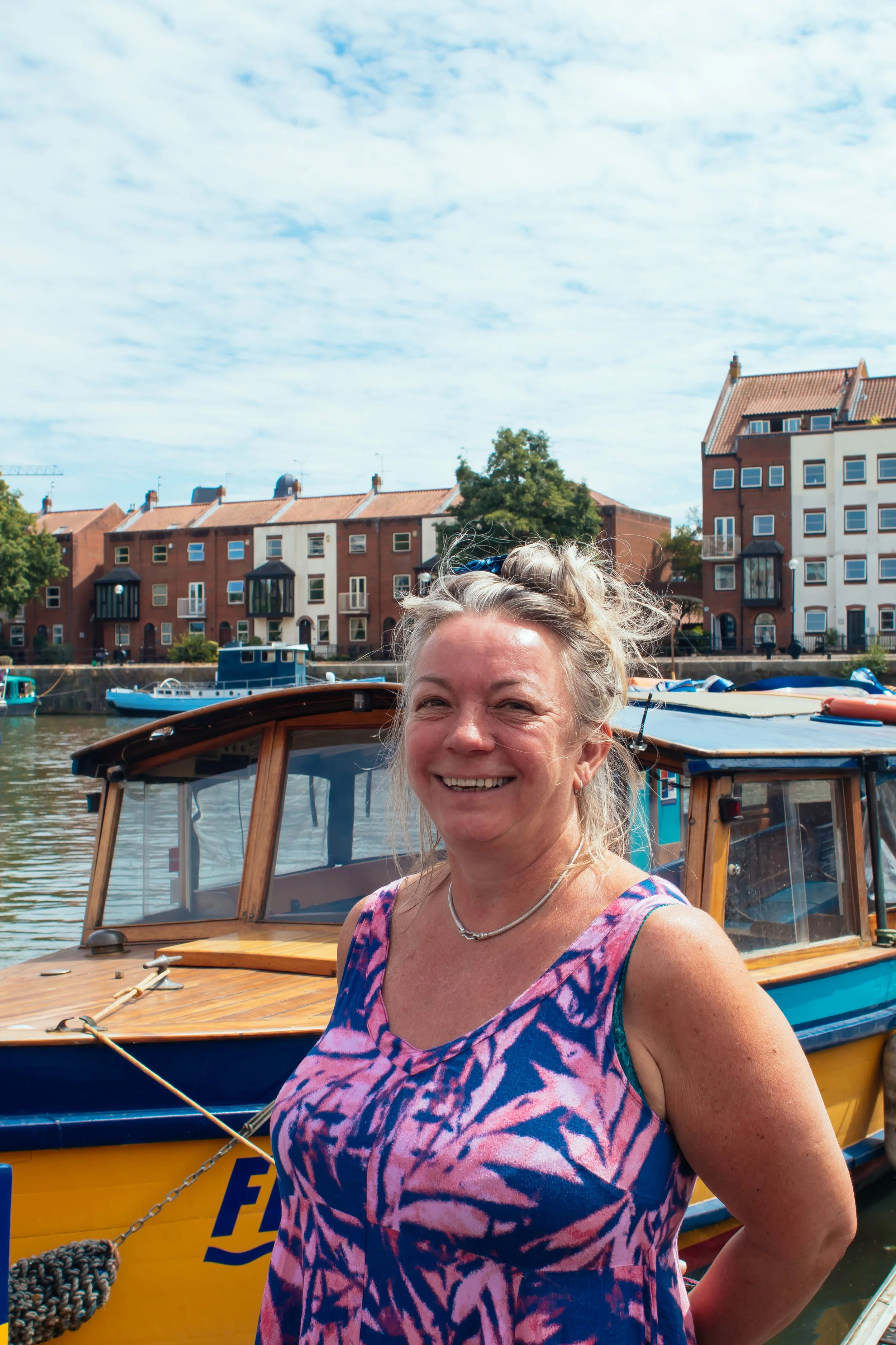 A woman with gray hair styled in a messy bun, smiling, standing in front of a boat on a body of water, with colorful buildings and a cloudy sky in the background.
