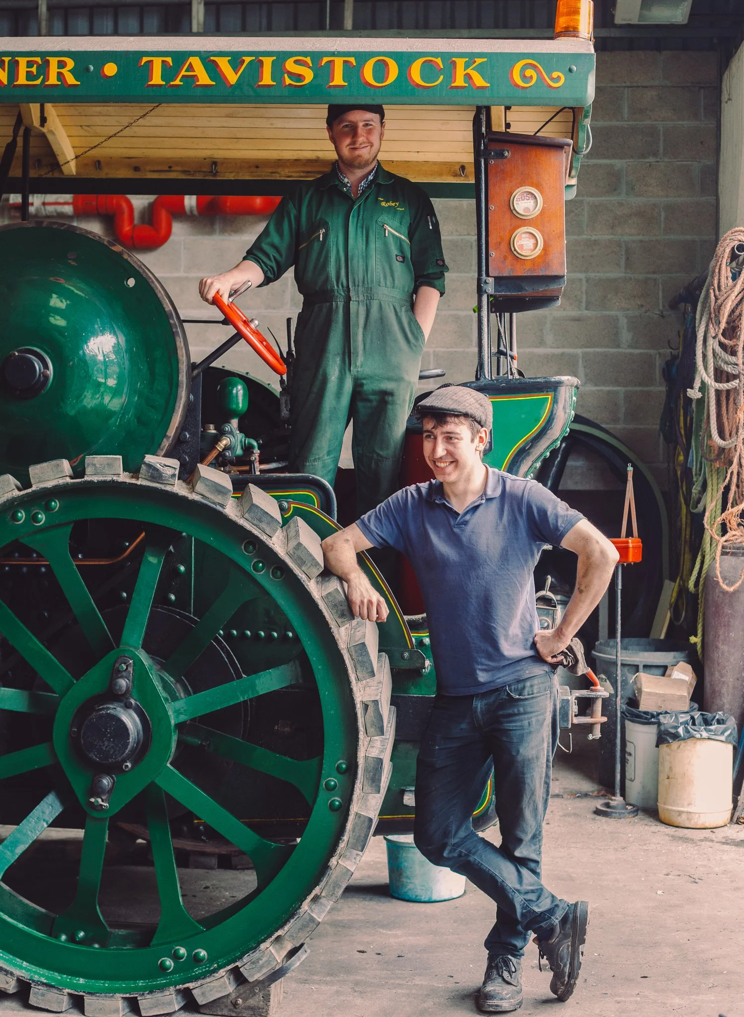Two men are posing with a vintage green steamroller inside a workshop. One man is standing on top of the machine, wearing green coveralls and a cap, holding onto the steering wheel. The other man is leaning against the large wheel, wearing a gray t-s