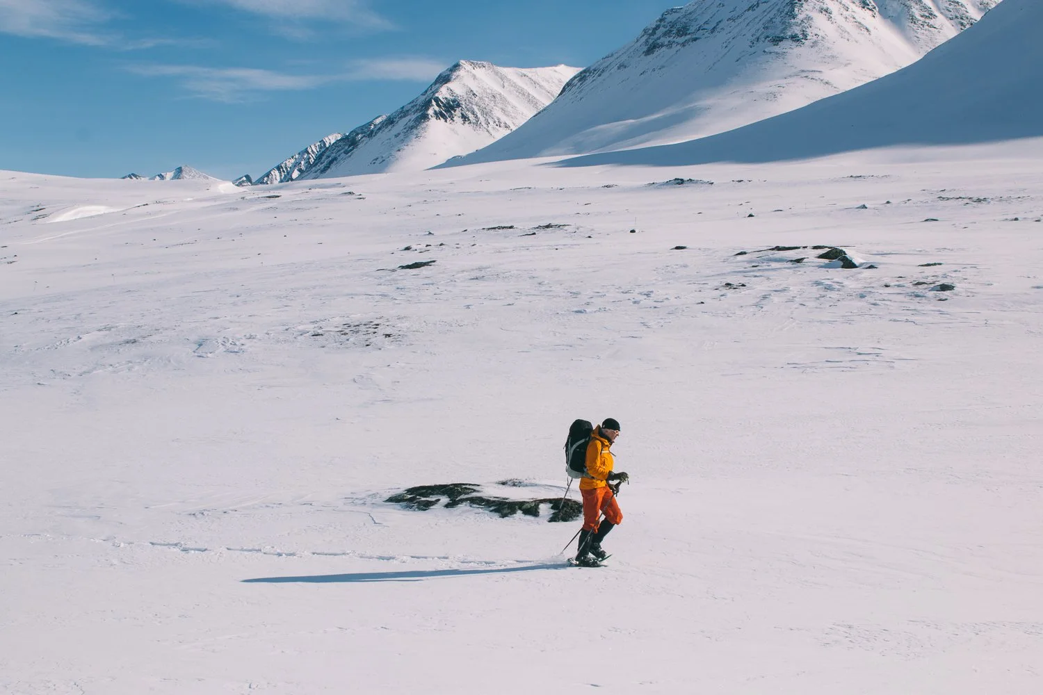 A person in an orange jacket and red pants cross-country skiing in a snowy landscape with mountains in the background.