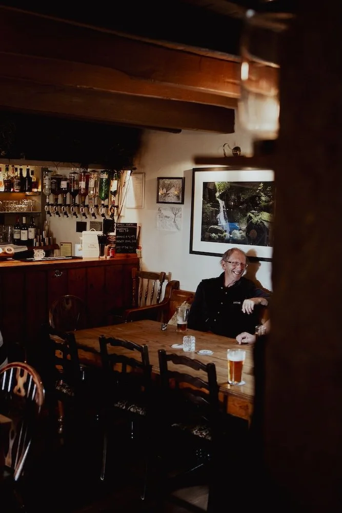 A man with glasses and a black shirt sitting at a long wooden table in a cozy bar or pub, smiling and drinking a beer, with glasses and a bottle on the table and a bar area with bottles and glasses in the background, along with framed pictures on the